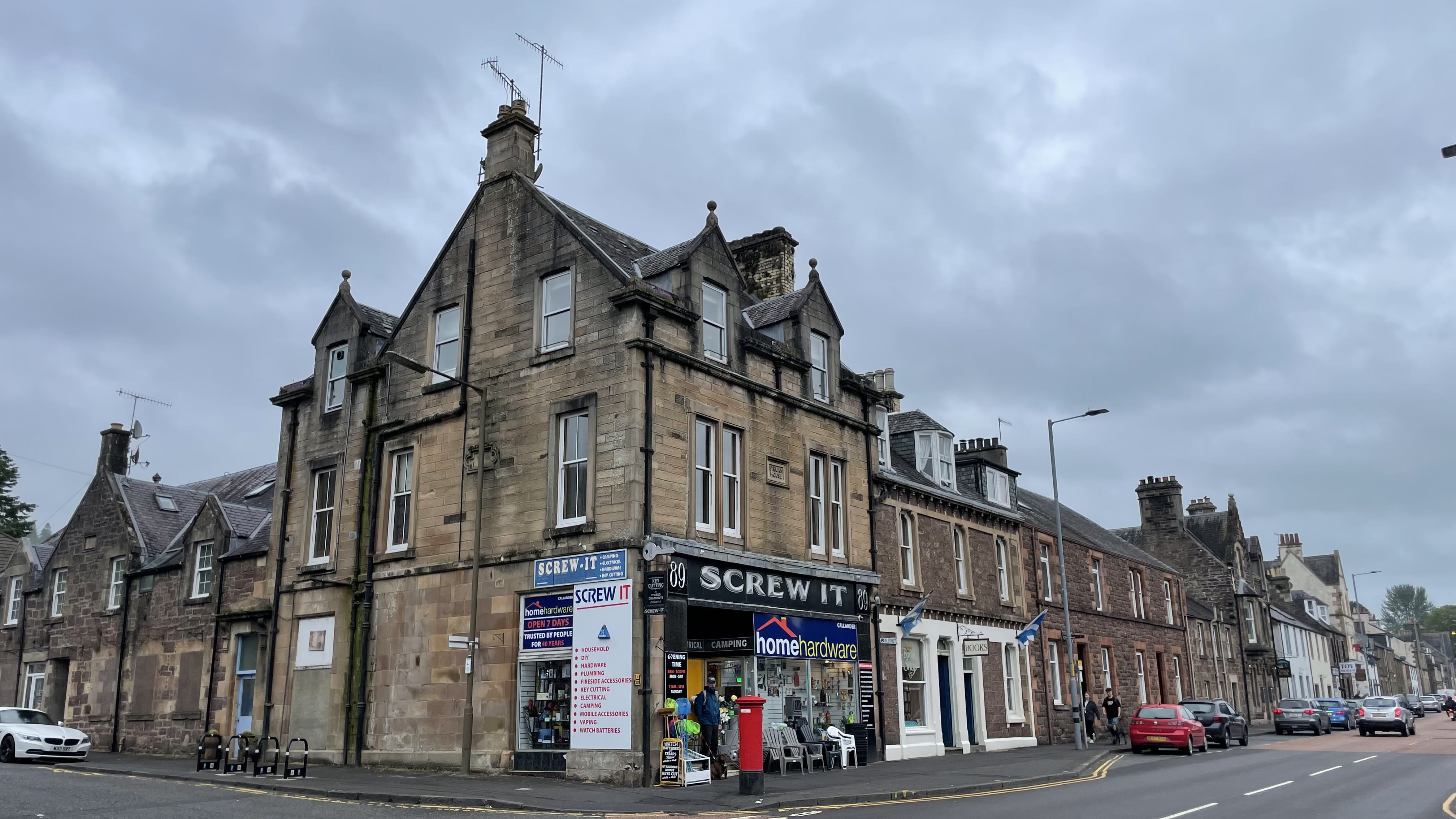 Stone building on Main Street in Callander, Scotland, with the corner hardware shop “Screw It” under a cloudy sky -Rob Roy Way