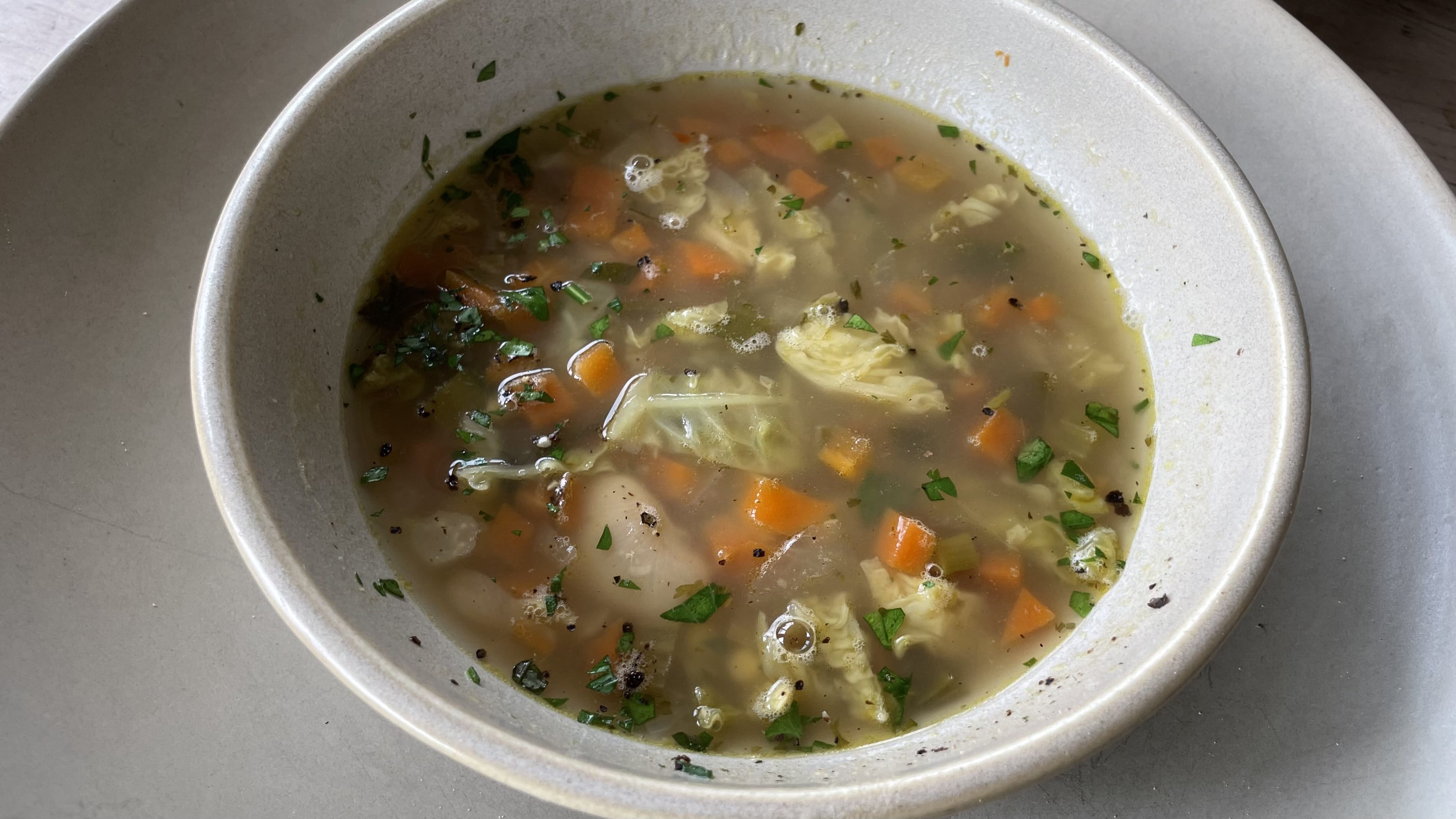 Bowl of vegetable soup with carrots, cabbage, and herbs served on a neutral-toned plate.