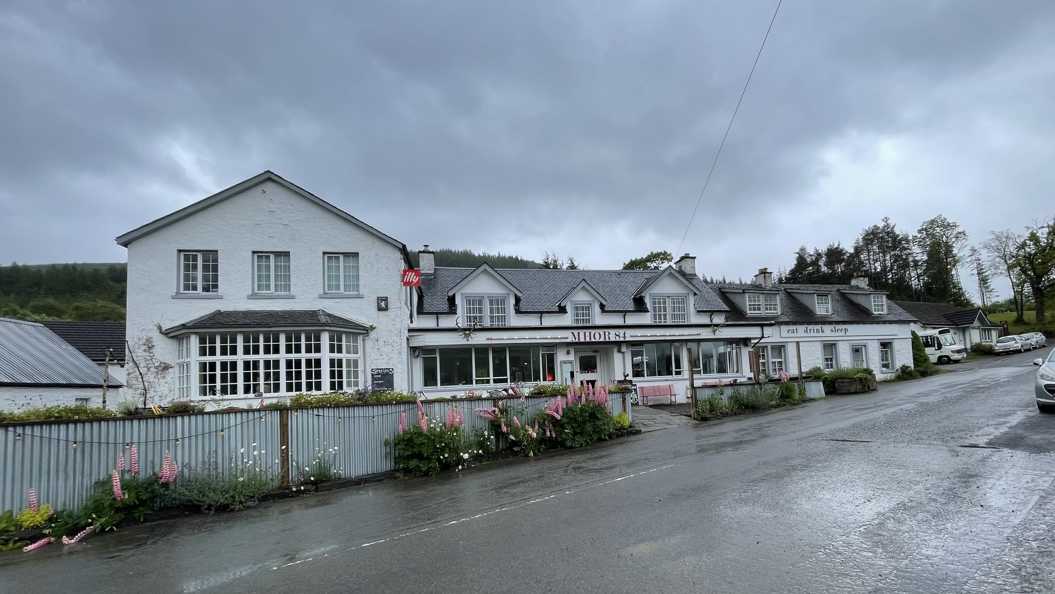 Mhor 84 roadside inn near Lochearnhead, Scotland, with whitewashed stone exterior and lupines blooming out front on a rainy day.