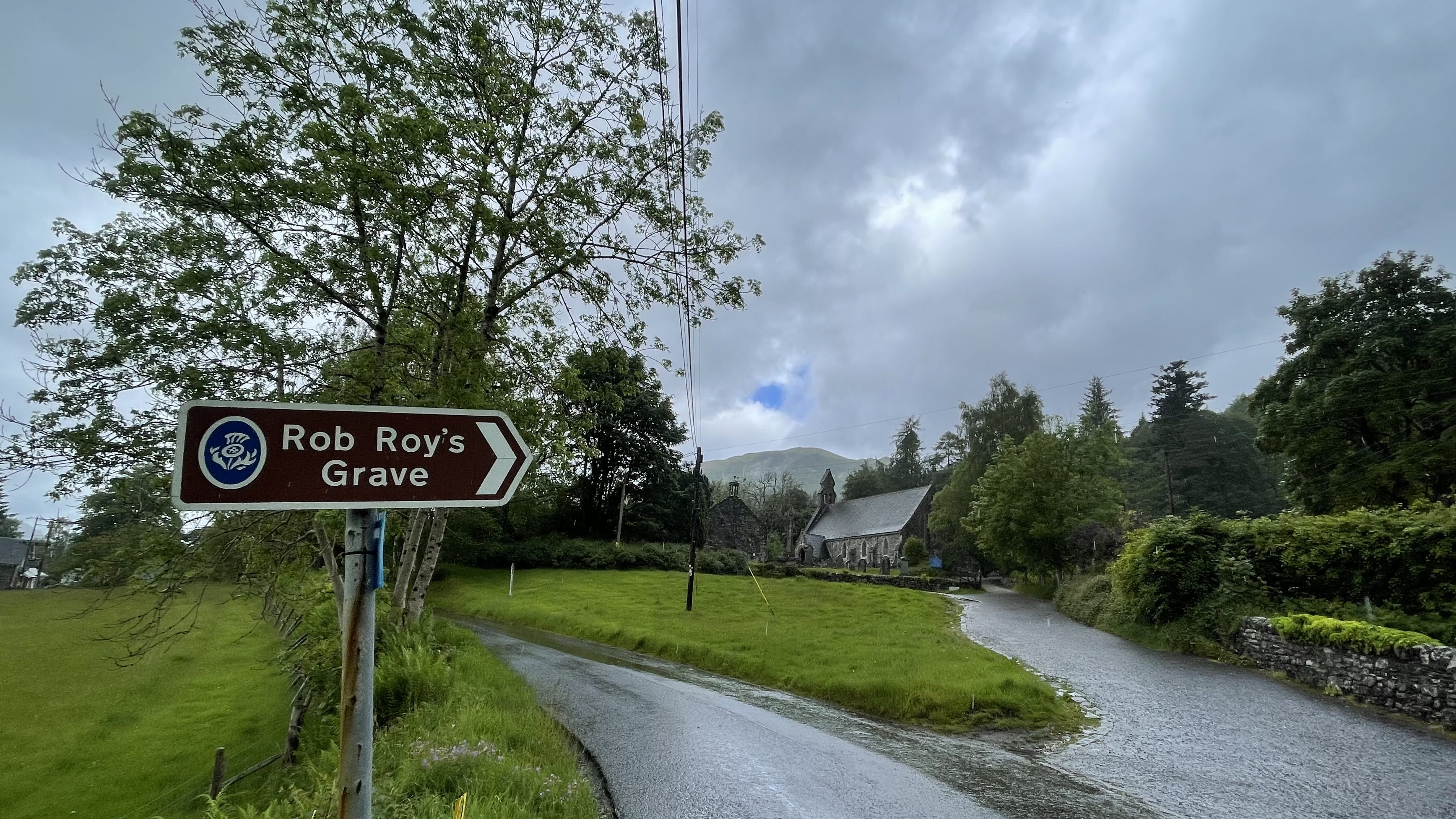 Road sign pointing to Rob Roy’s Grave along a wet country road in Balquhidder, Scotland, with a stone church and green hills under a cloudy sky
