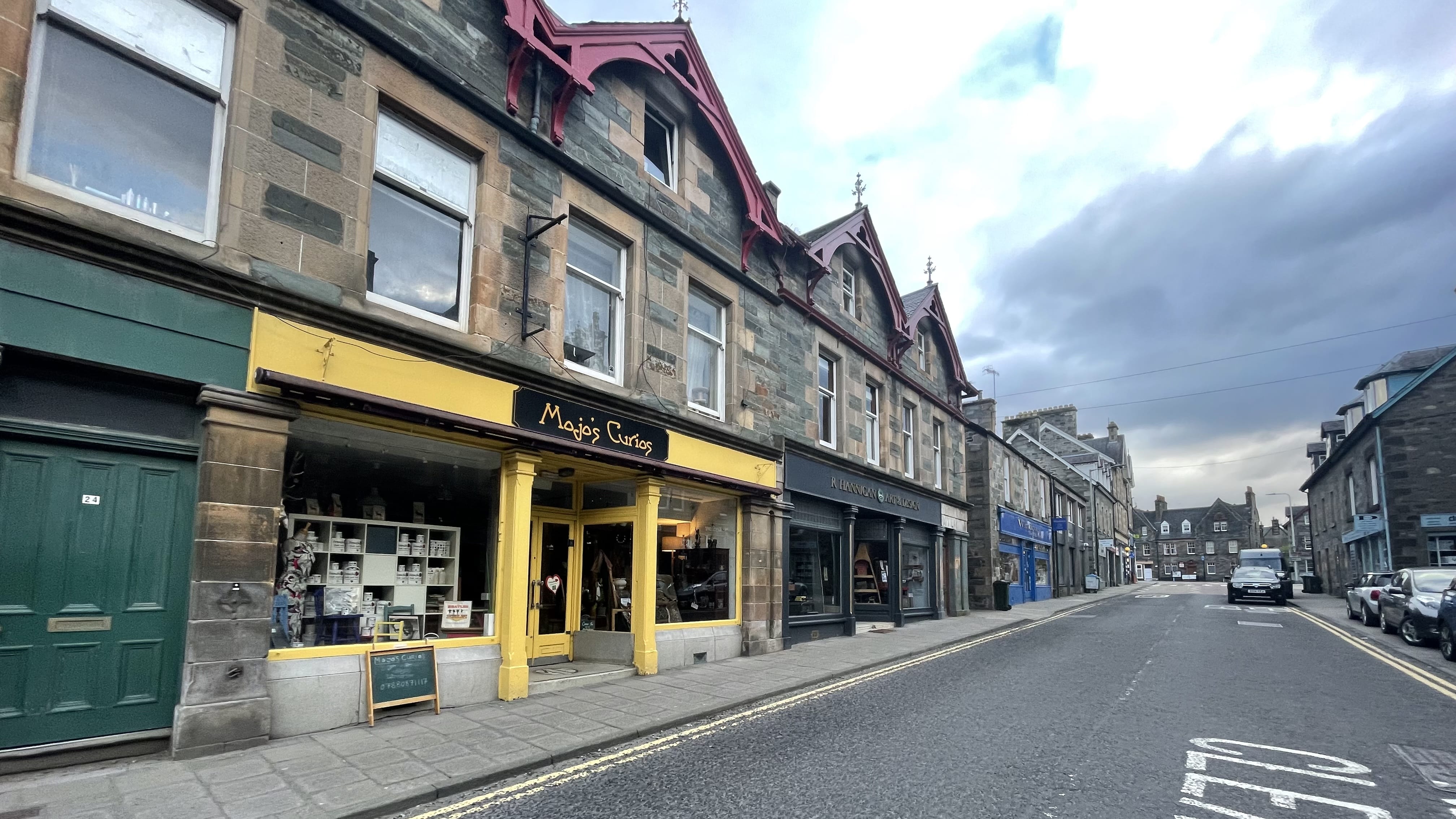Colorful storefronts along a quiet street in Aberfeldy, Scotland, featuring a yellow shopfront called Mojo’s Curios and traditional stone buildings under a cloudy sky