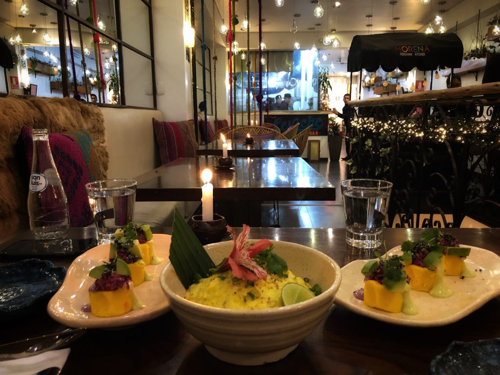Table at Morena Peruvian Kitchen in Cusco, Peru, set with elegant dishes including colorful causa rolls and a quinoa-based entrée, surrounded by candles, woven pillows, and soft lighting.