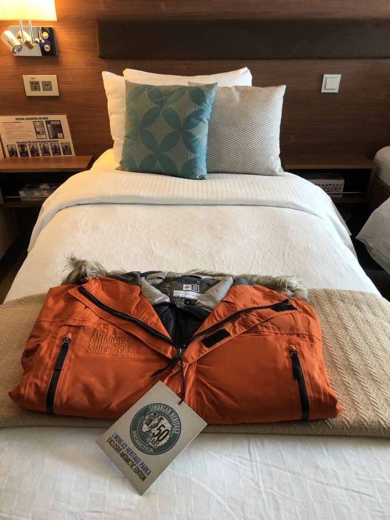 Bright orange Lindblad Expeditions Antarctica parka laid out on a neatly made bed in a cabin aboard the National Geographic Explorer, with a commemorative 50th anniversary tag.