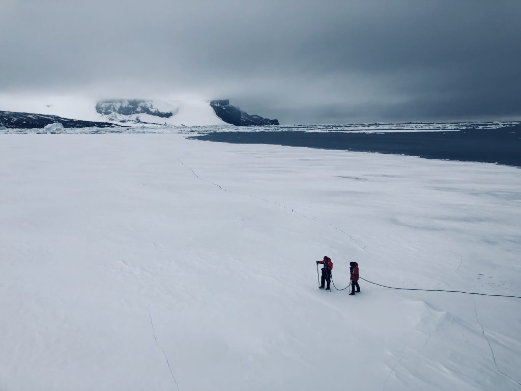 Two red-jacketed expedition staff walking across sea ice near James Ross Island, Antarctica, roped together under a dramatic overcast sky.