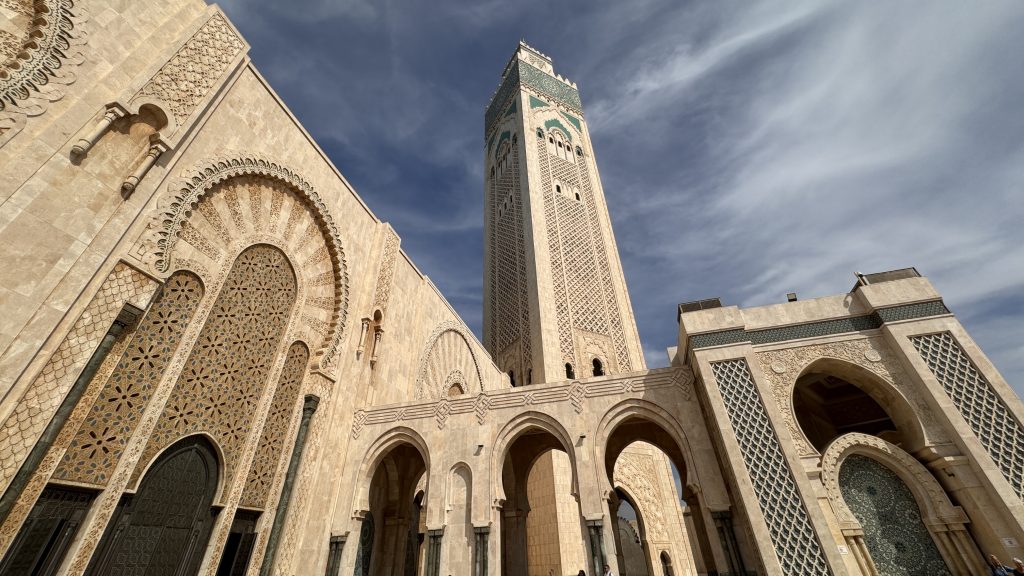 Low-angle view of the Hassan II Mosque in Casablanca, Morocco, showing the ornate arches, carved stonework, and towering minaret against a blue sky.