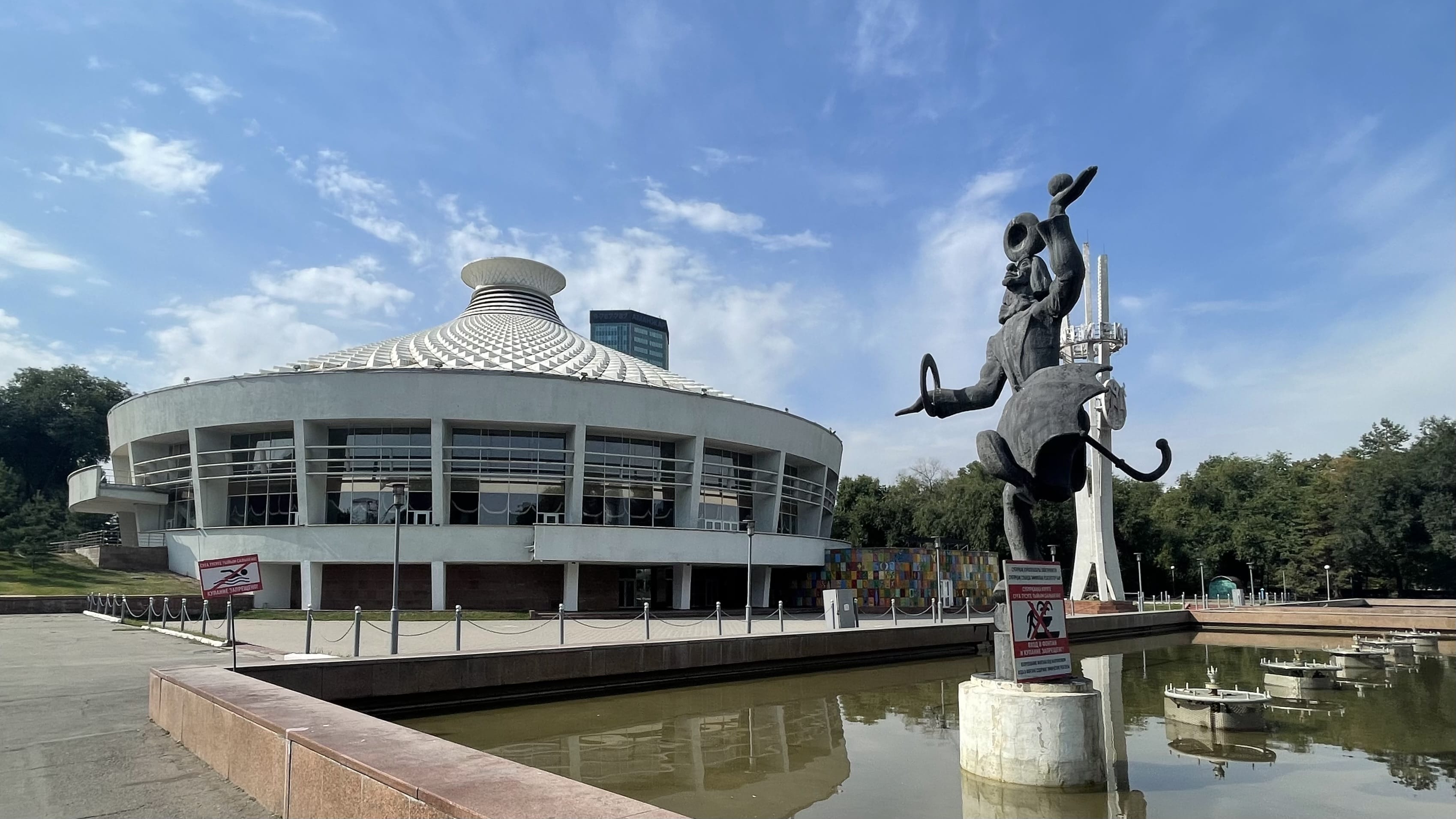 Almaty Circus in Kazakhstan, a circular Soviet-era building with a domed roof and a clown statue standing in a reflecting pool under a blue sky