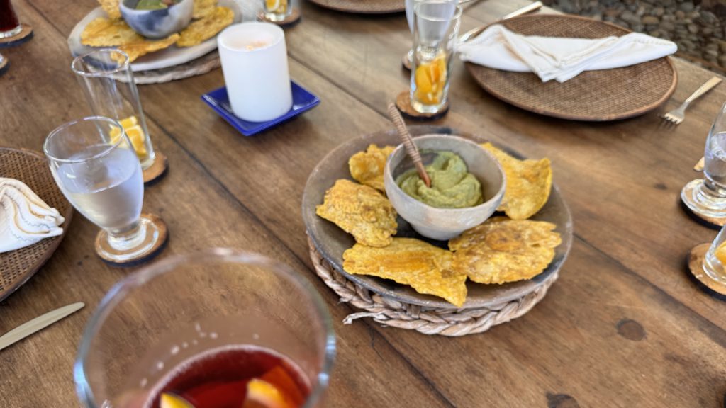 A wooden table set for lunch with glasses of sangria and water, woven placemats, and a centerpiece of crispy plantain chips served with guacamole in a clay bowl.