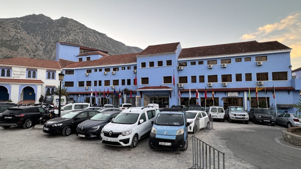 2 Days in Chefchaouen. A large hotel building painted bright blue with terracotta tile roofing, multiple windows, and Moroccan architectural details, set against a mountainous backdrop at sunset.