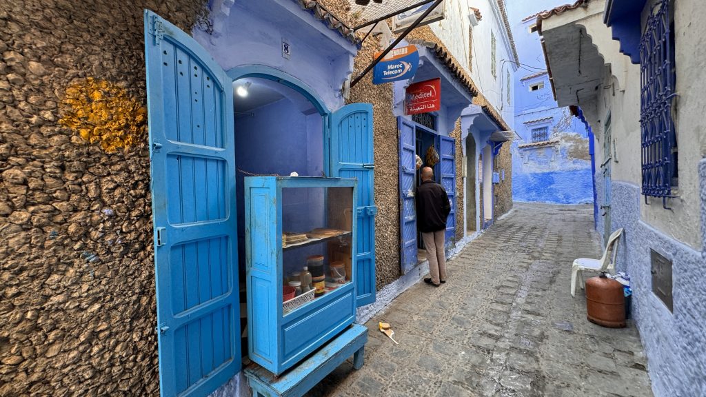 Small blue-painted shop in Chefchaouen with open wooden doors and a display case showing jars and flatbreads, along a quiet cobblestone street.
