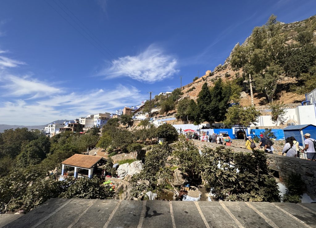 View of Ras El Ma spring area in Chefchaouen, Morocco, with people walking along the stone path above the natural spring and wash station, nestled below the hills and whitewashed buildings under a clear blue sky.