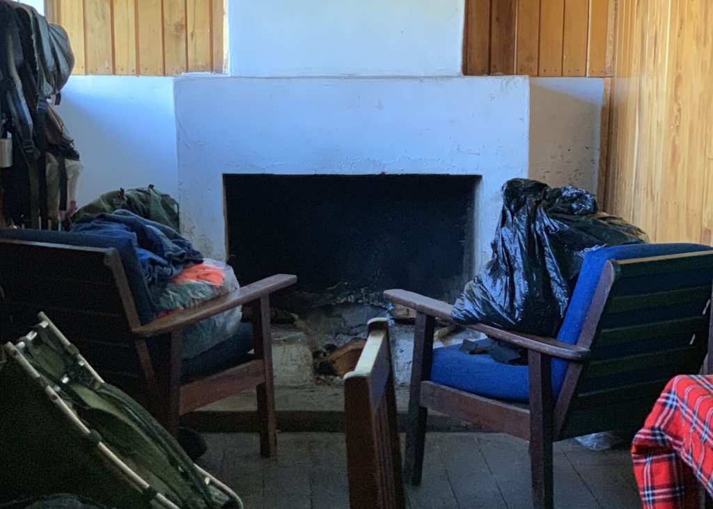 Simple sitting area inside Meru Mount Kenya Bandas with wooden chairs, a fireplace, and hikers’ gear piled nearby.