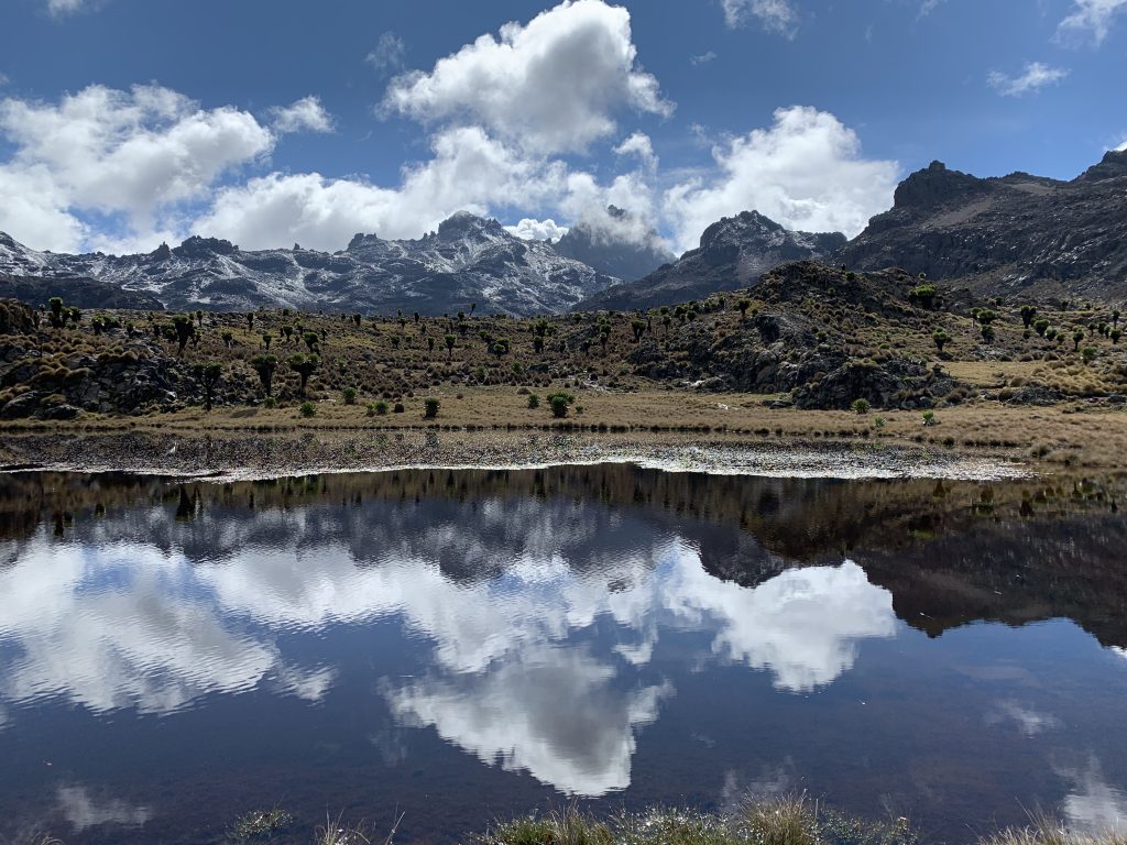 Alpine lake at Mintos Camp on Mount Kenya, reflecting jagged snow-dusted peaks and clouds in the still water.