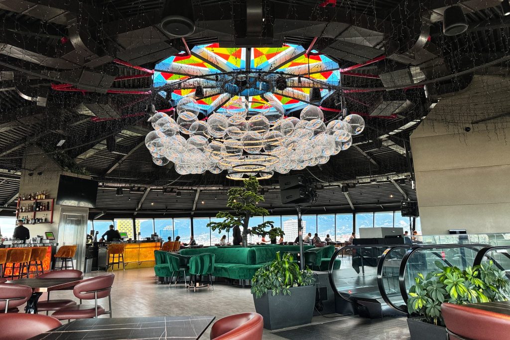 Interior of the revolving Sky Bar at the Sky Hotel in Tirana, featuring a colorful stained-glass ceiling, hanging glass bubble lights, and panoramic city views.