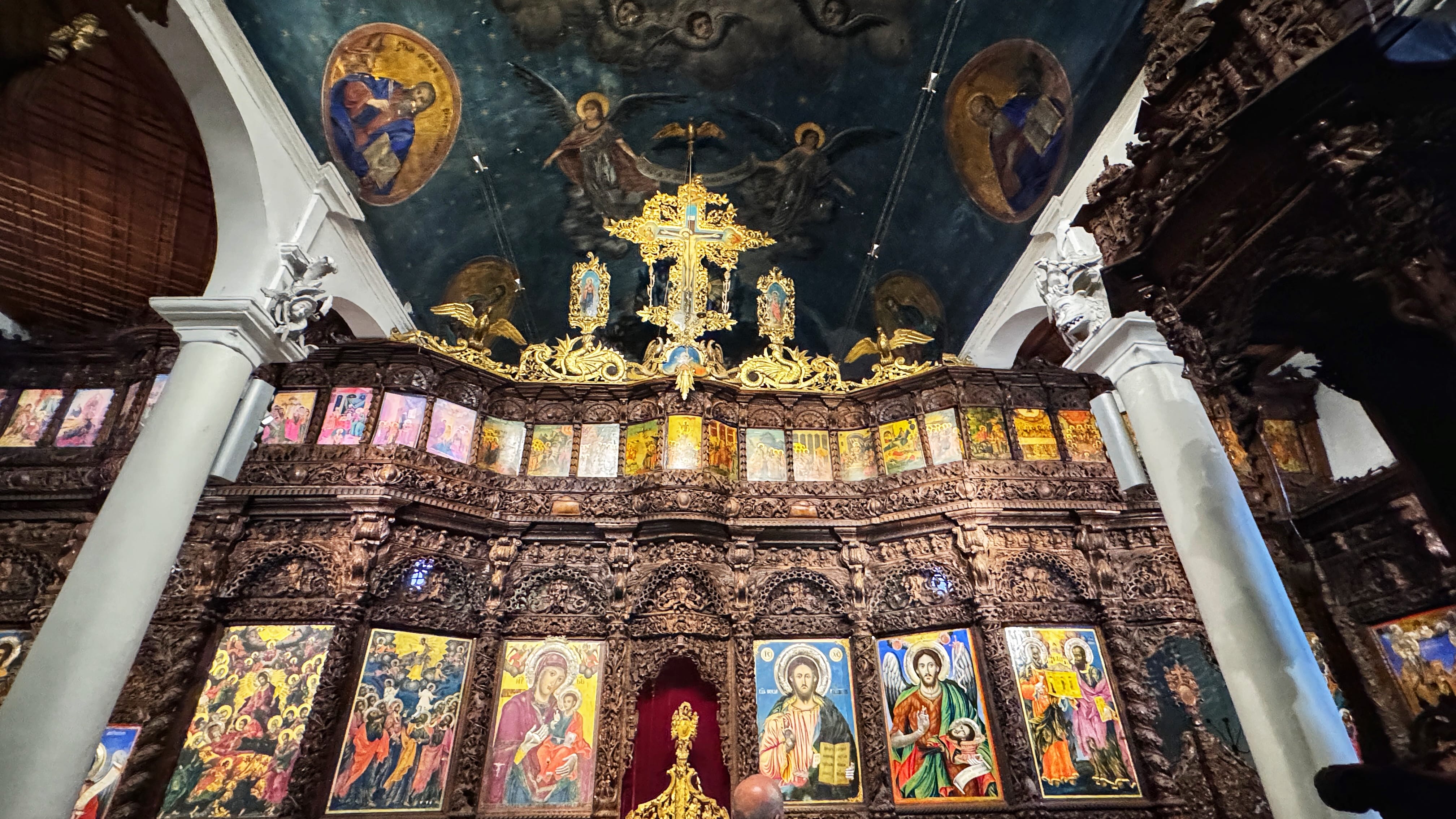 Ornate carved wooden iconostasis inside a Macedonian Orthodox church in Skopje, filled with colorful religious icons and crowned with a gold cross and celestial ceiling paintings.