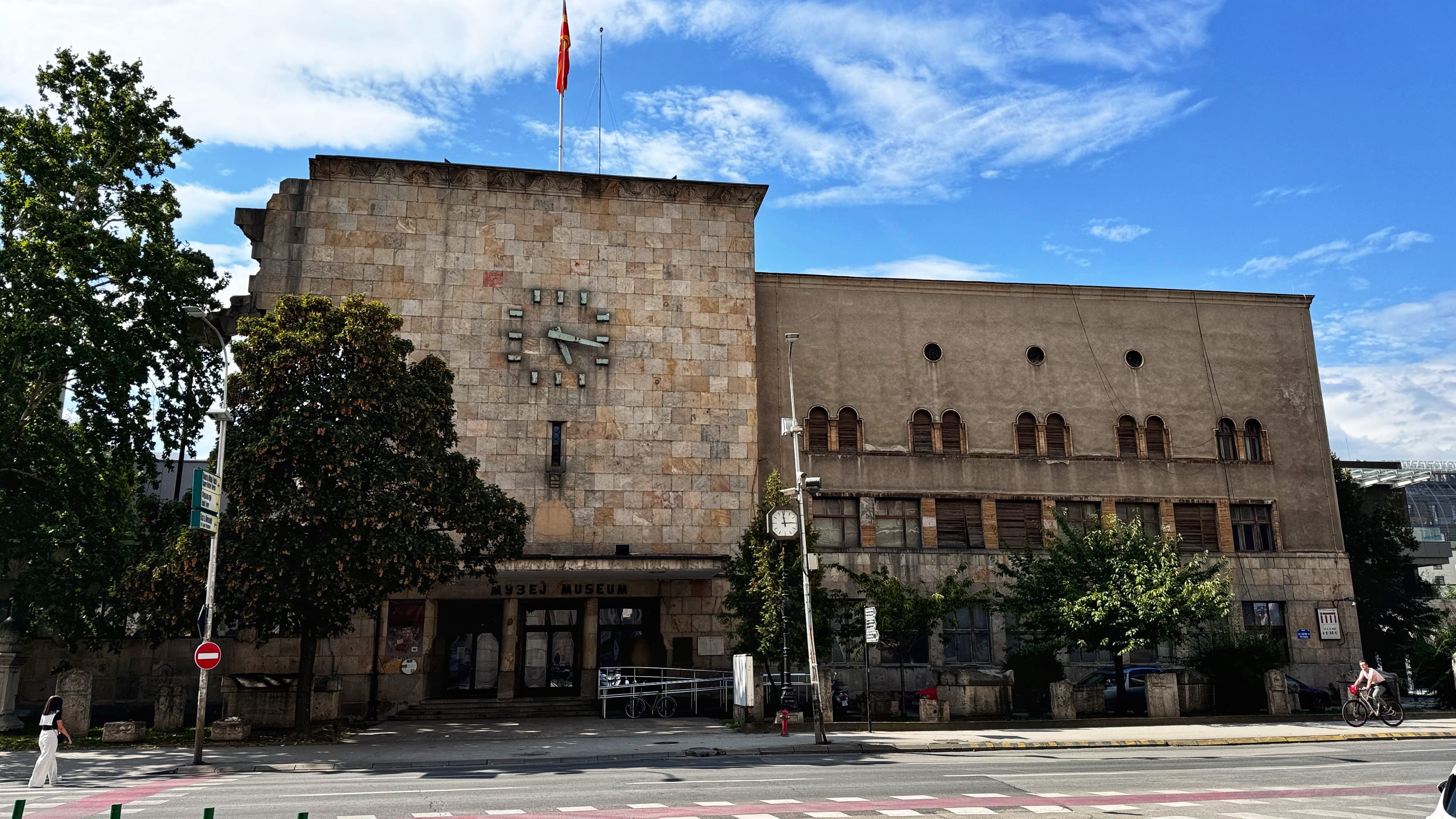 A stone building in Skopje, North Macedonia, with a damaged left facade and a large clock frozen at 5:17. The structure houses the Museum of the City of Skopje and was formerly the city’s railway station, partially destroyed during the 1963 earthquake.