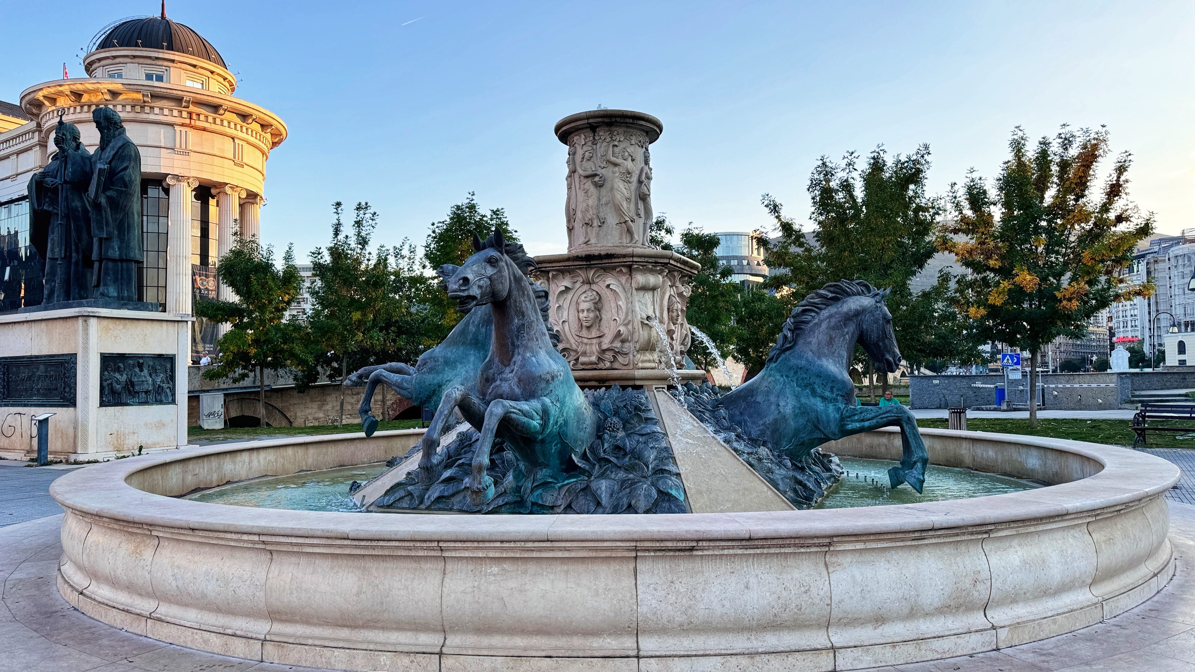 A decorative fountain in Skopje, North Macedonia, featuring dramatic bronze horse sculptures emerging from water among sculpted leaves. Behind them is a central stone column adorned with classical figures and reliefs. To the left, another monument depicts two robed men, possibly historical scholars or saints. A neoclassical domed building is visible in the background.