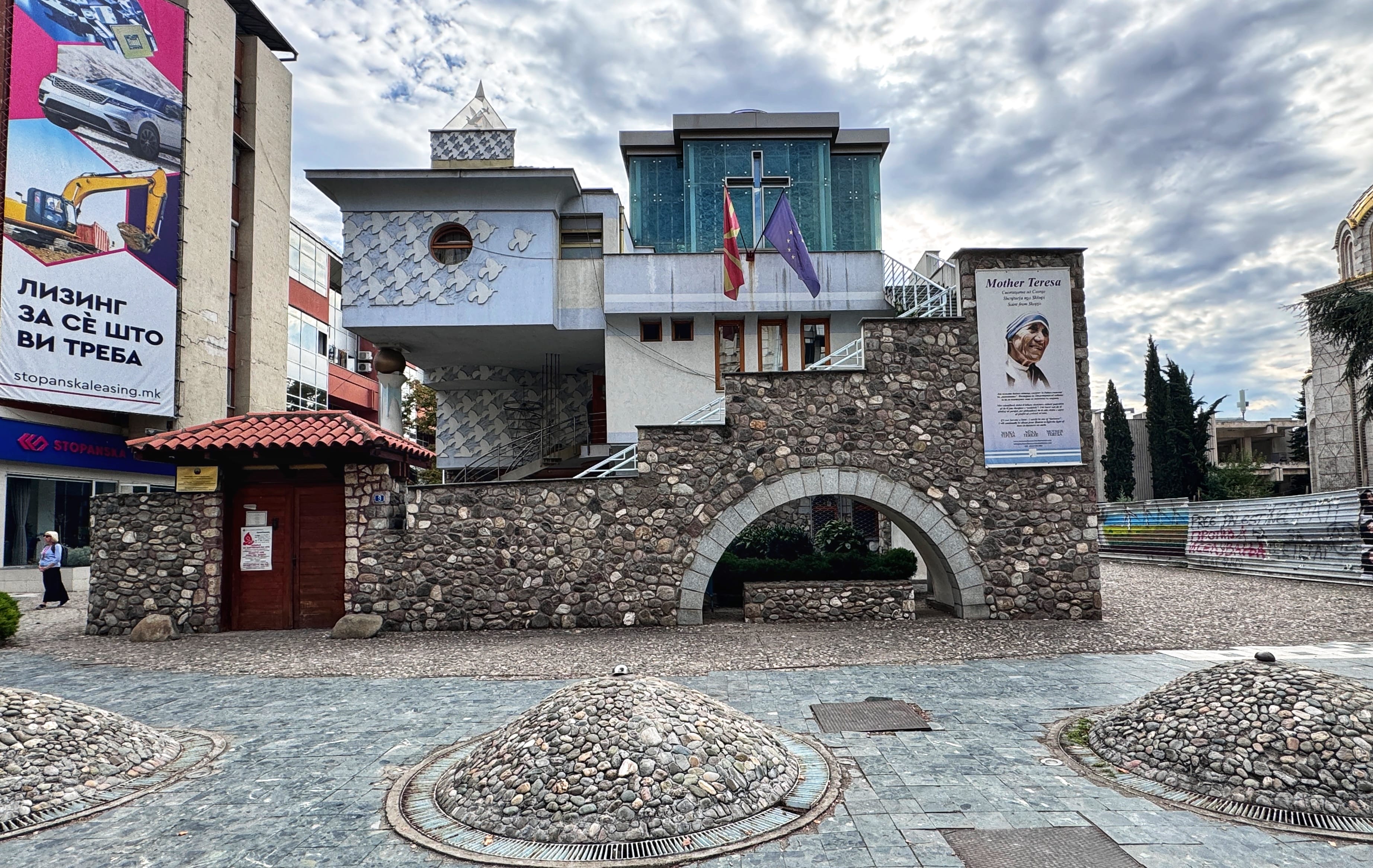 The Mother Teresa Memorial House in Skopje, North Macedonia, featuring modern architecture with butterfly motifs, stone walls, and a banner honoring Mother Teresa.