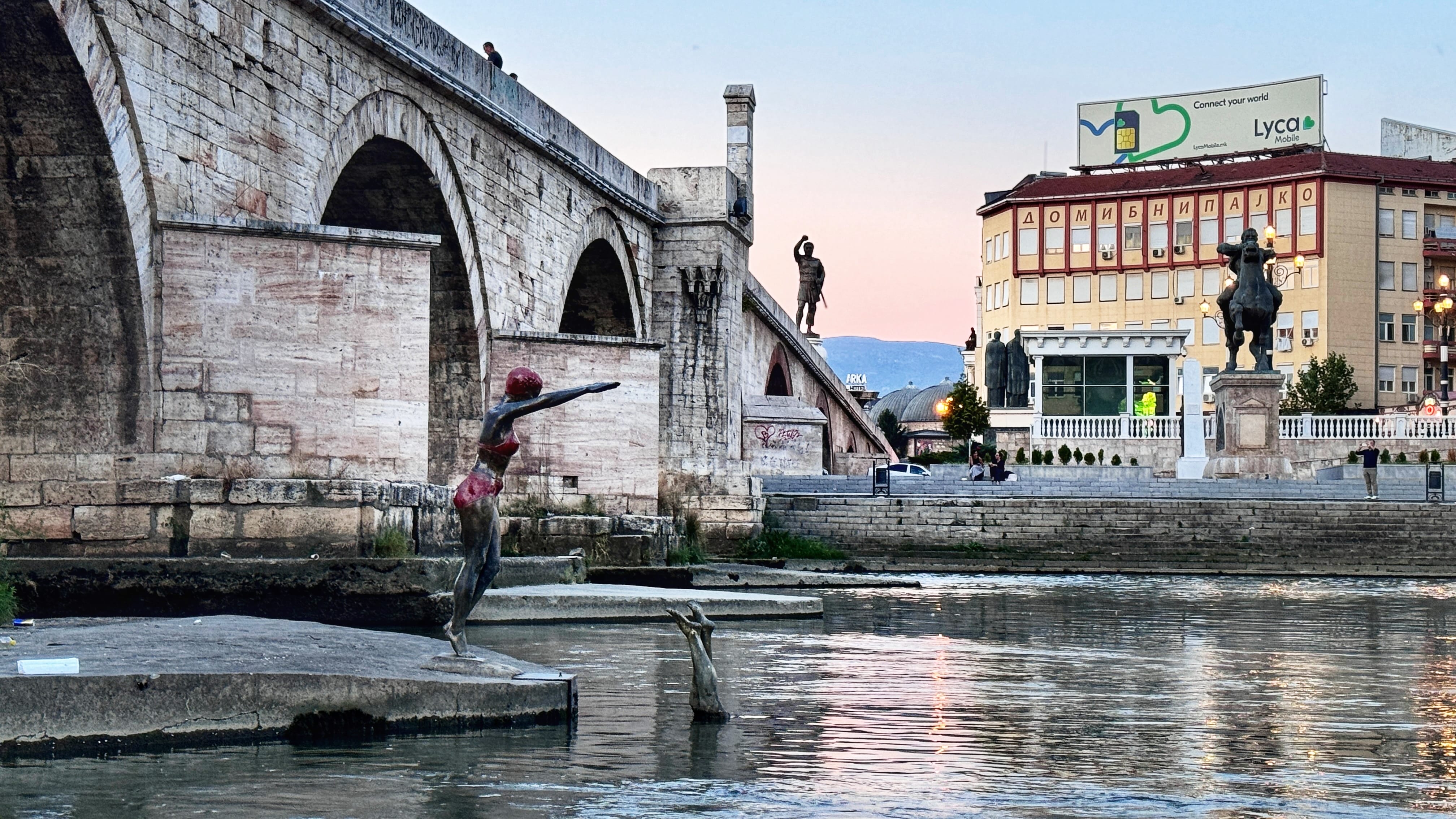 View of the Stone Bridge in Skopje, North Macedonia, crossing the Vardar River at sunset. In the foreground, a bronze statue of a swimmer in a red bathing cap and suit prepares to dive. Across the bridge, additional statues—including an equestrian figure—stand in Macedonia Square, with city buildings and mountains in the background.