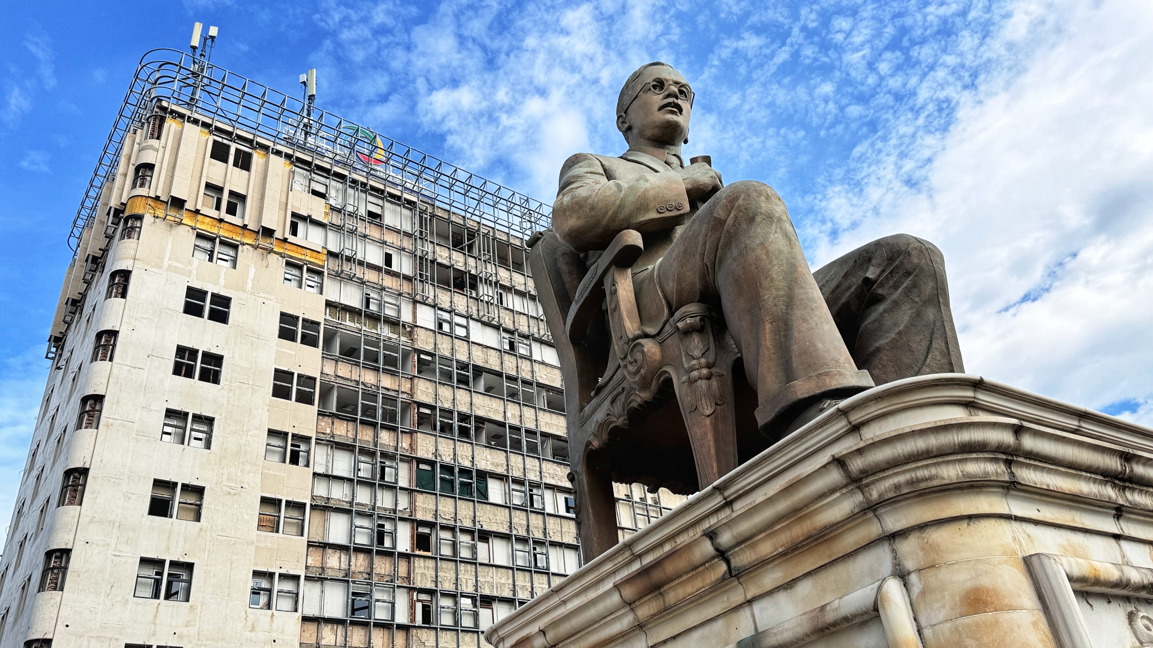 A large bronze statue of a man seated in an ornate chair dominates the foreground, set against a dilapidated high-rise building in Skopje, North Macedonia. The building features exposed concrete, stripped facade panels, and a visible metal framework, contrasting sharply with the polished monument. The juxtaposition highlights the city’s blend of monumental ambition and post-socialist decay.