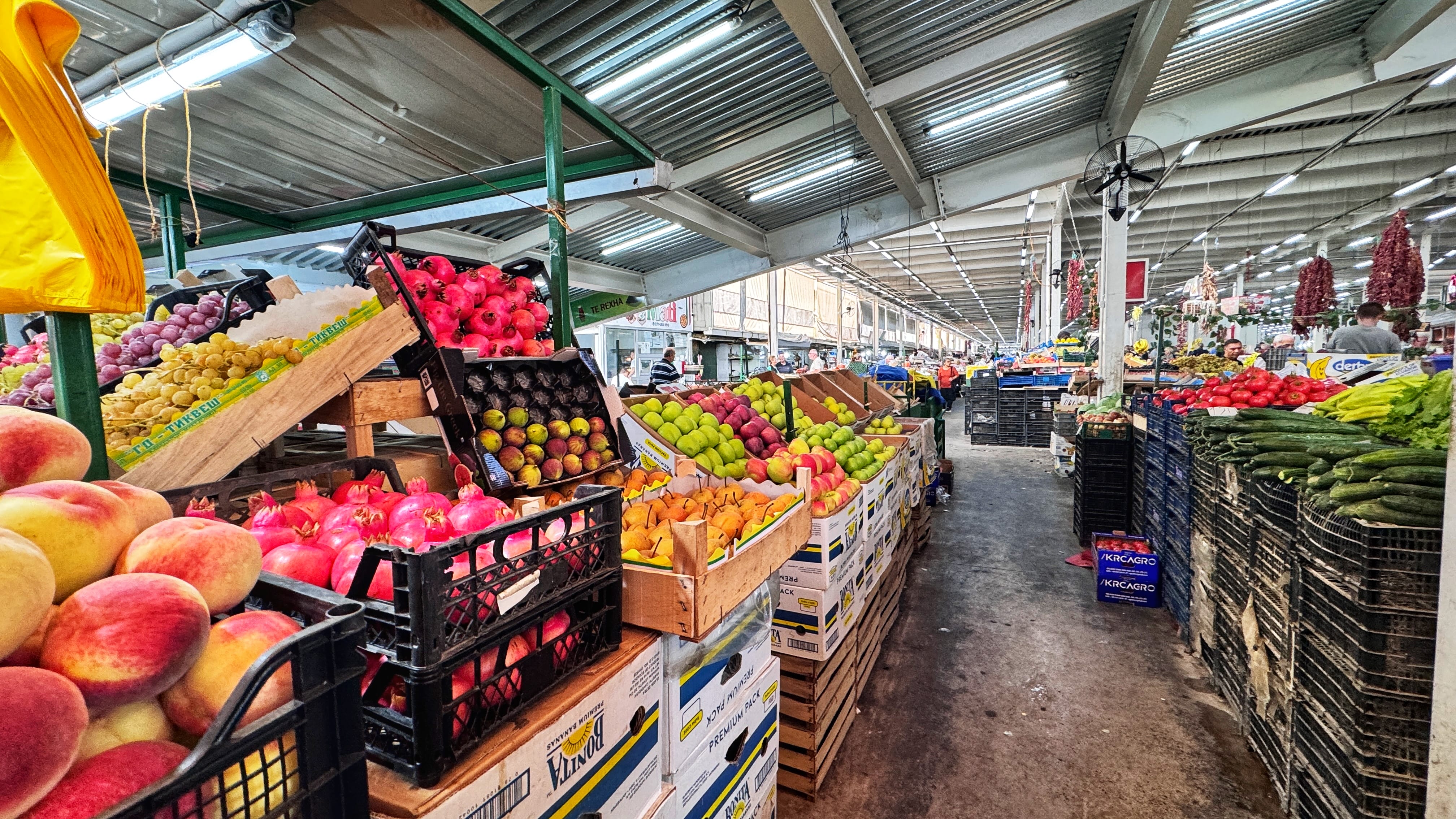 A colorful open-air fruit market stall in Skopje filled with pears, apples, bananas, grapes, and citrus fruits. A vendor stands nearby, chatting with customers.