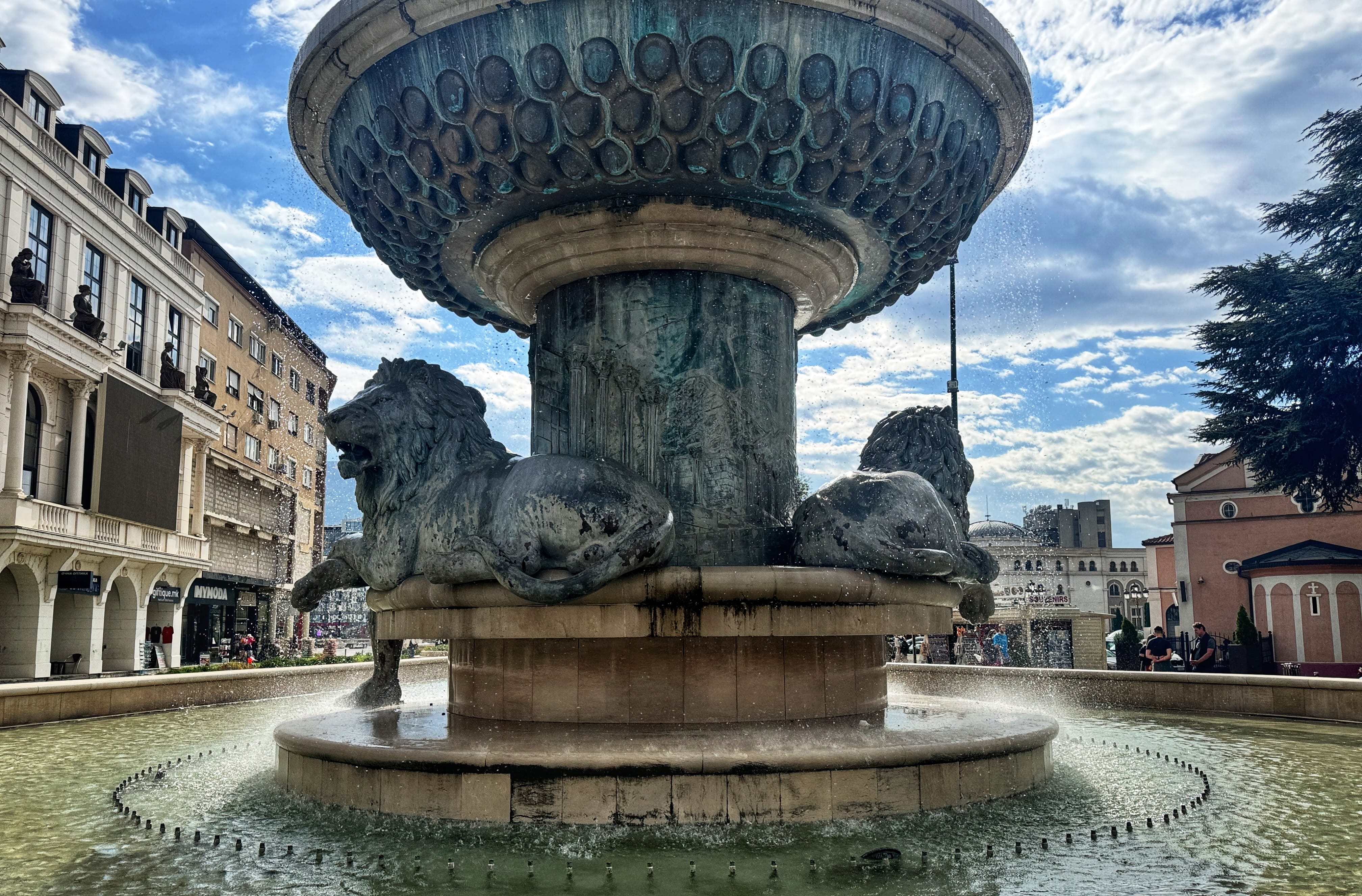 A close-up view of bronze lion sculptures resting around the base of a large, ornate fountain in Skopje’s city center. Water flows from multiple levels of the structure, with classic-style buildings and a partly cloudy sky in the background.