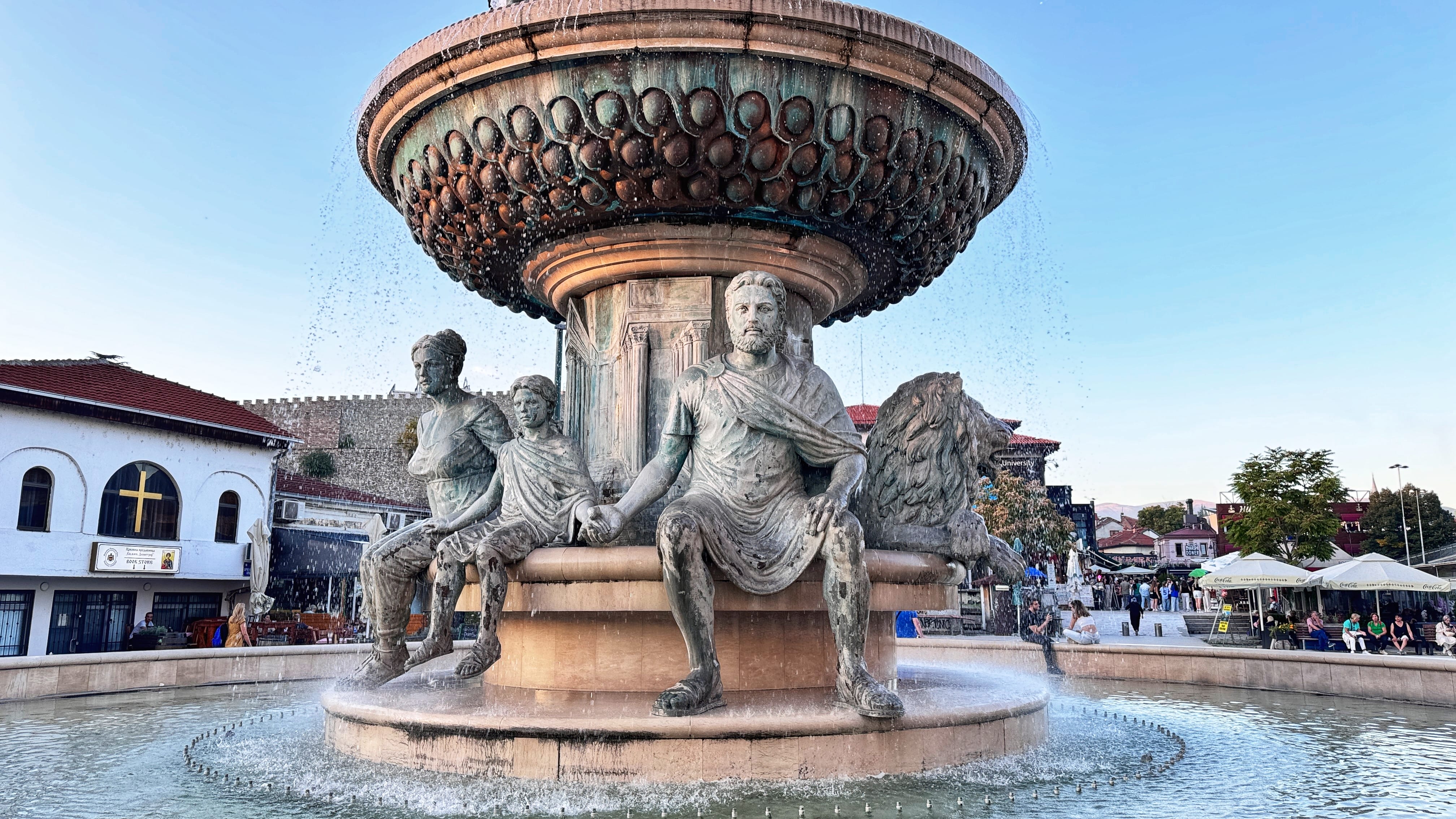 A neoclassical-style fountain in Skopje featuring bronze sculptures of a seated man, woman, and child holding hands, flanked by lions. Water flows from an ornate upper bowl, with shops, a church, and fortress walls visible in the background.