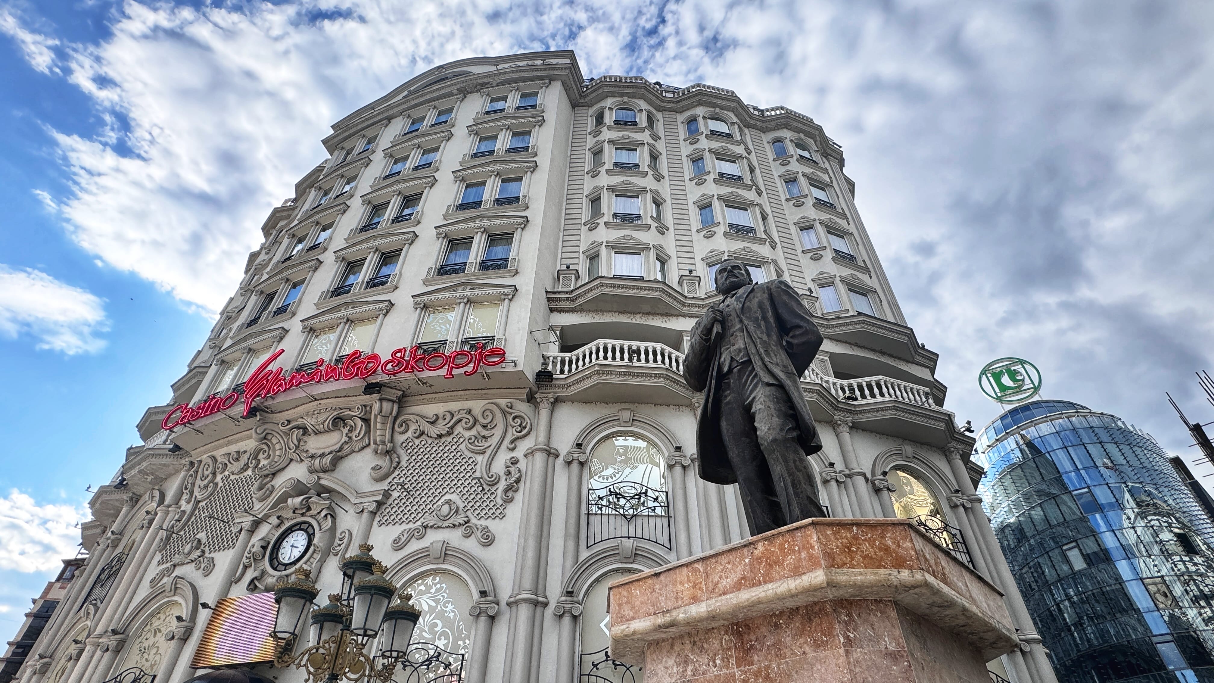 A low-angle view of a neoclassical-style building in Skopje, North Macedonia, with ornate architectural details and a neon sign reading "Casino Flamingo Skopje." In the foreground stands a bronze statue of a man in formal attire. To the right, a modern glass building with a green logo contrasts the older structure.
