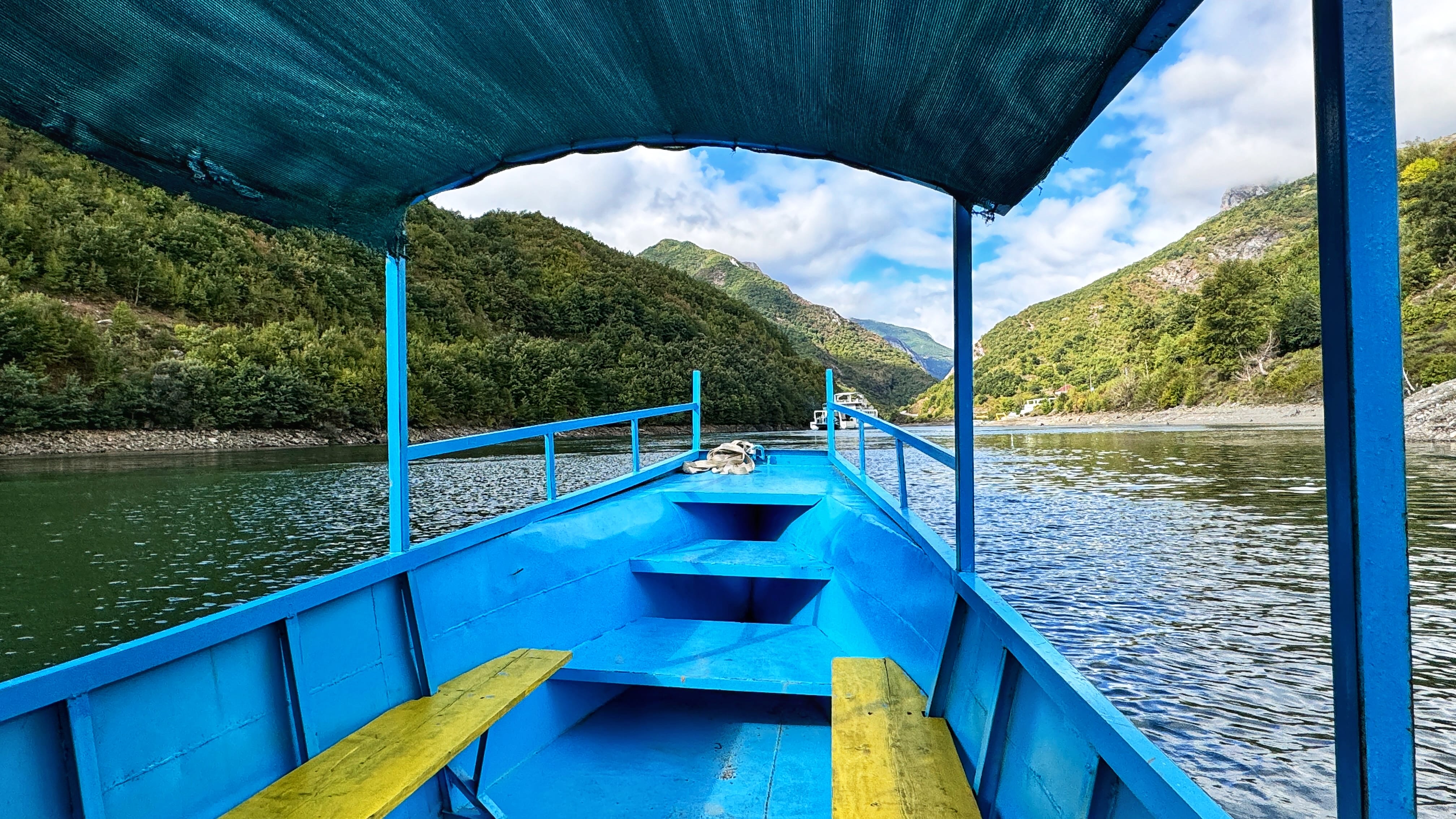 View from inside a bright blue boat with a canopy, cruising through the scenic green hills and calm waters of Komani Lake in Albania.