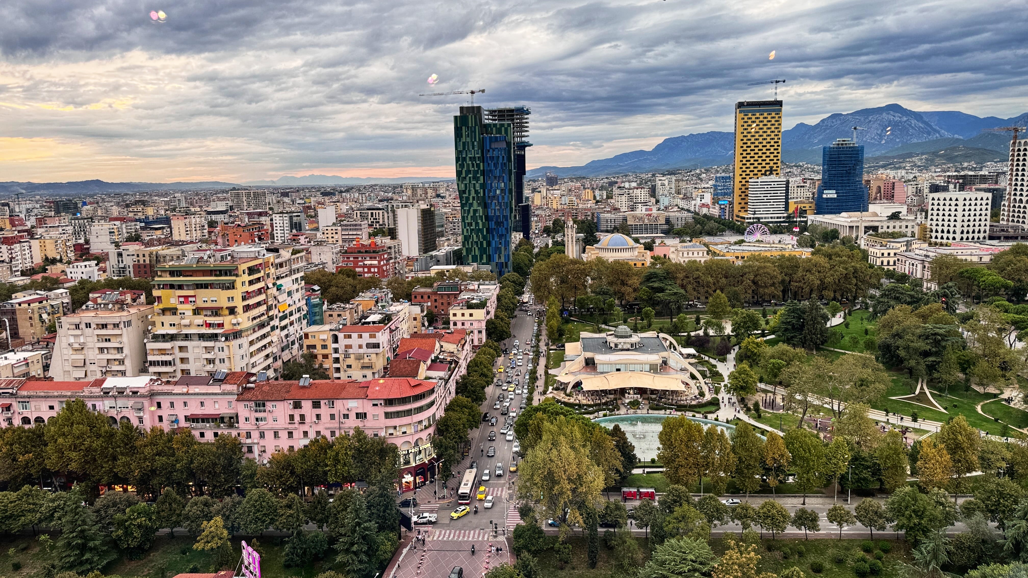 Aerial view of Tirana, Albania, showing colorful apartment blocks, tree-lined streets, and surrounding hills under a cloudy sky.