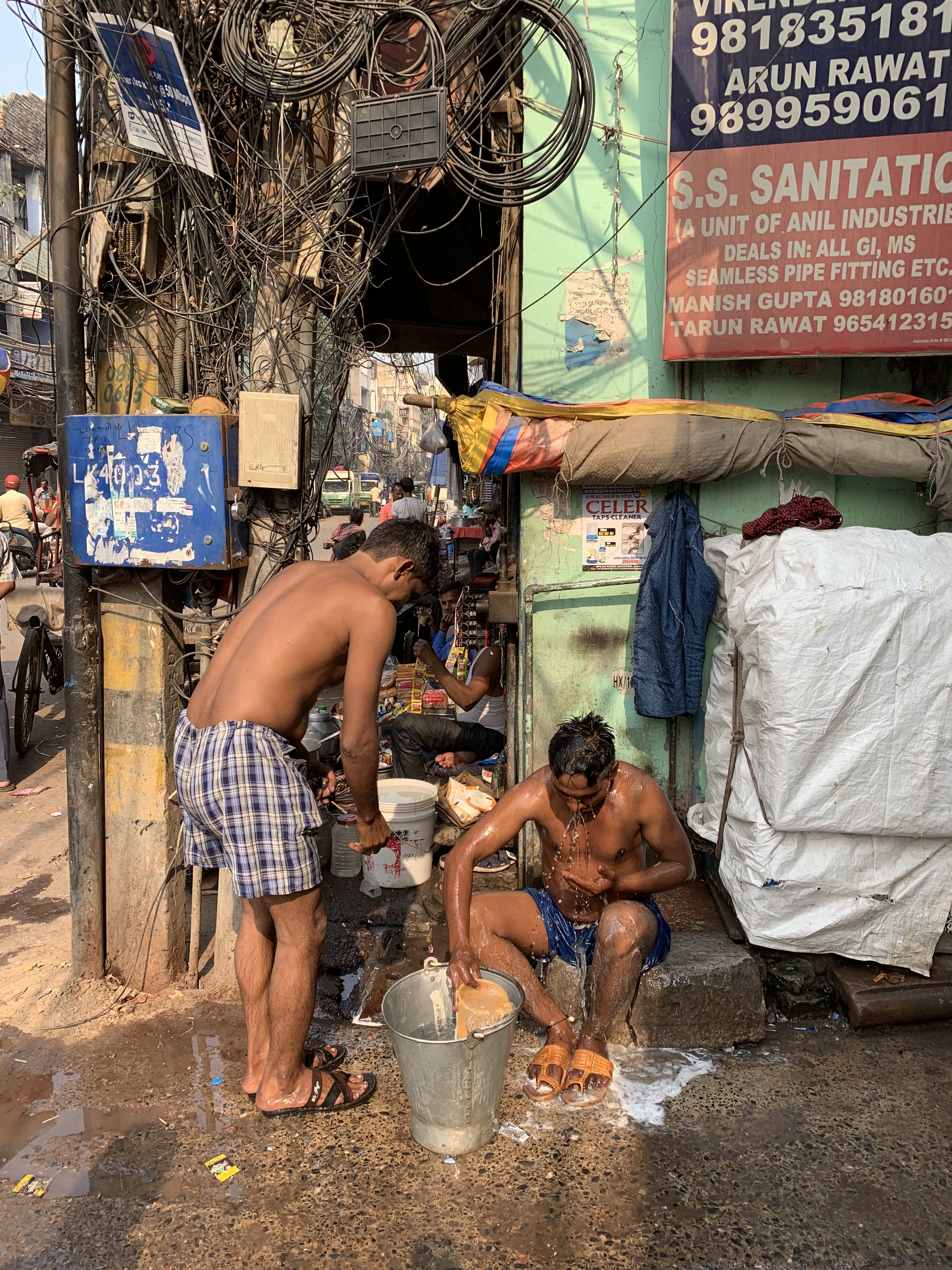 Two shirtless men bathing with buckets of water on a Delhi street corner surrounded by exposed wires.
