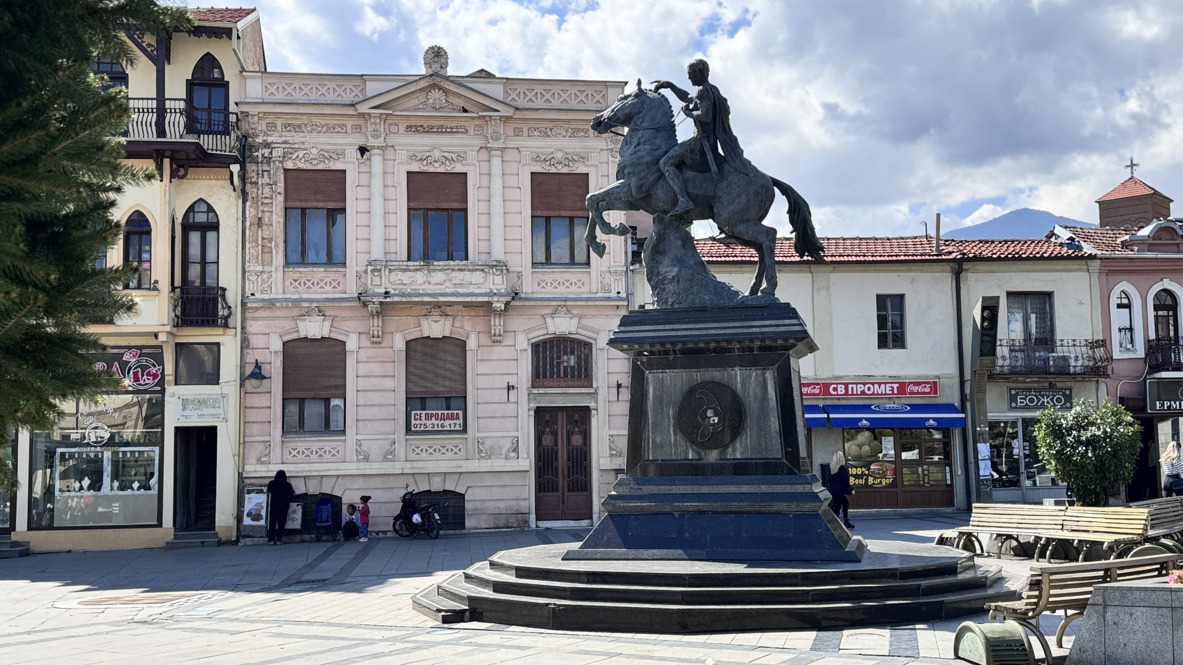 A statue of a rider on a rearing horse stands in the center of a square in Bitola, North Macedonia. The bronze equestrian monument is set on a dark stone pedestal, surrounded by benches and a mix of neoclassical and modest storefront buildings. Mountains rise in the distance under a partly cloudy sky.