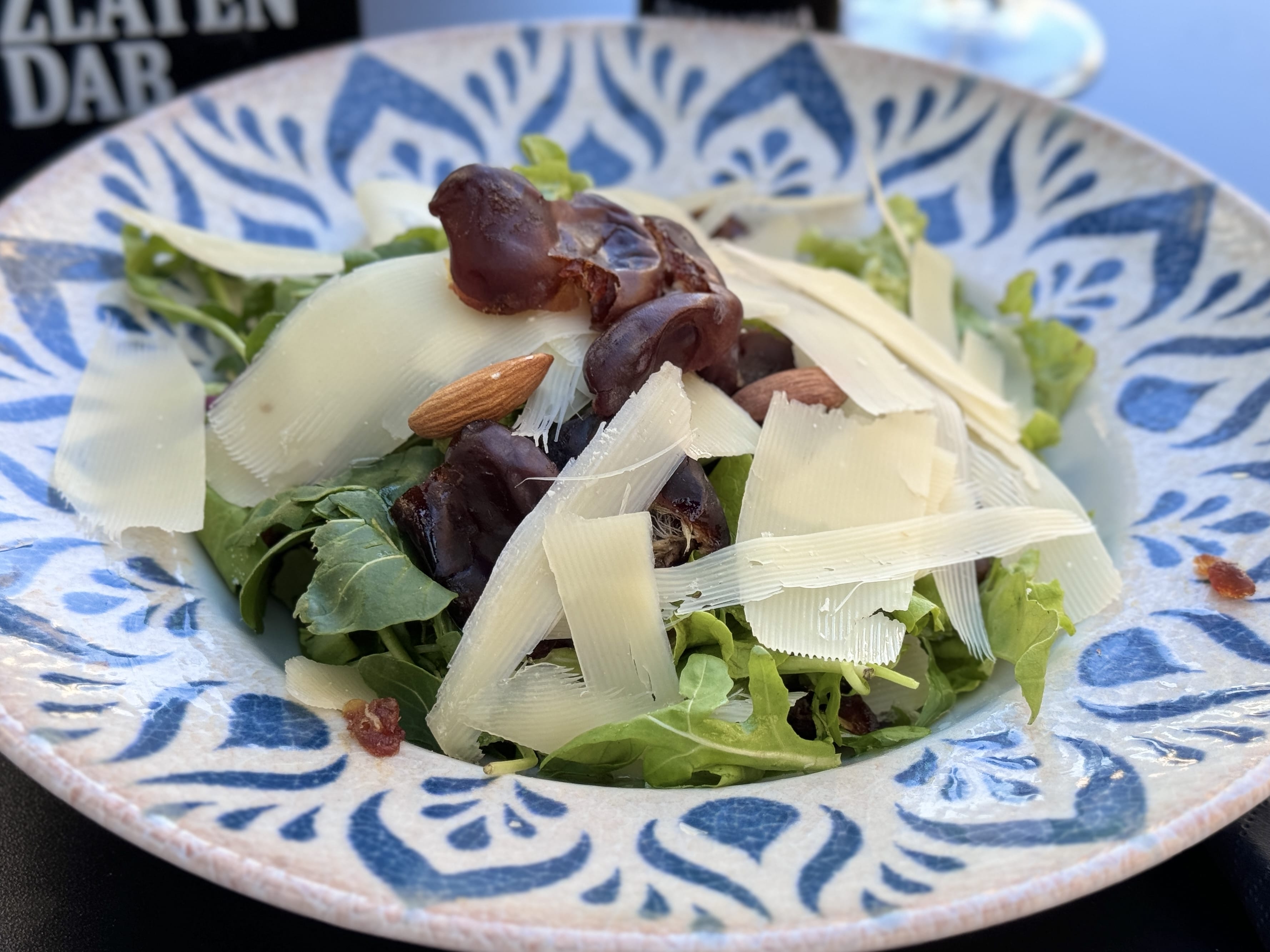 A fresh salad served in a decorative blue-and-white bowl, featuring leafy greens topped with thin shavings of cheese, halved dates, whole almonds, and a drizzle of dressing. The ingredients are vibrant and arranged naturally for an appealing presentation.