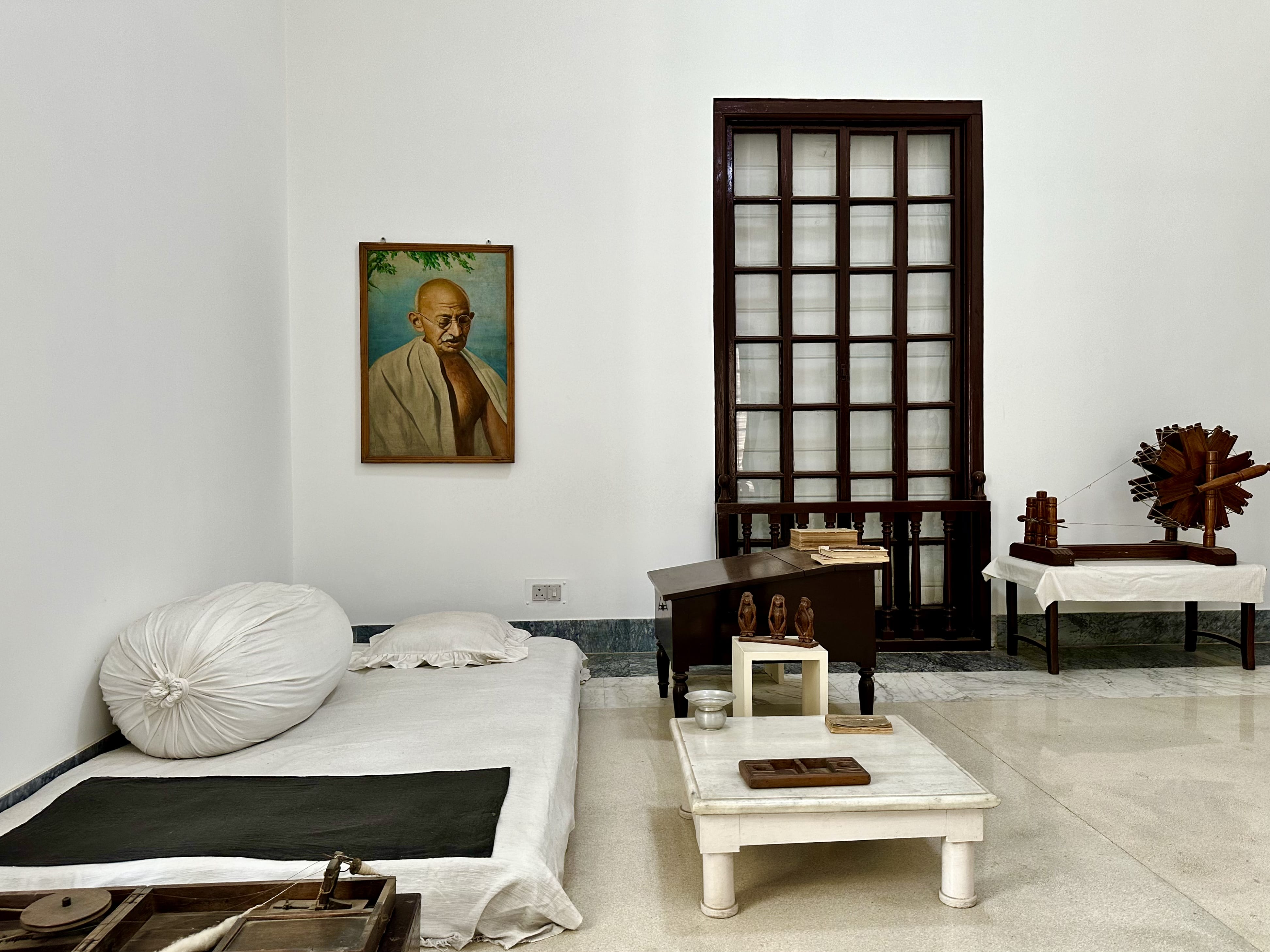 A simple room with Gandhi’s low bed, white cushions, spinning wheel, and personal items, with a portrait of Gandhi on the wall.