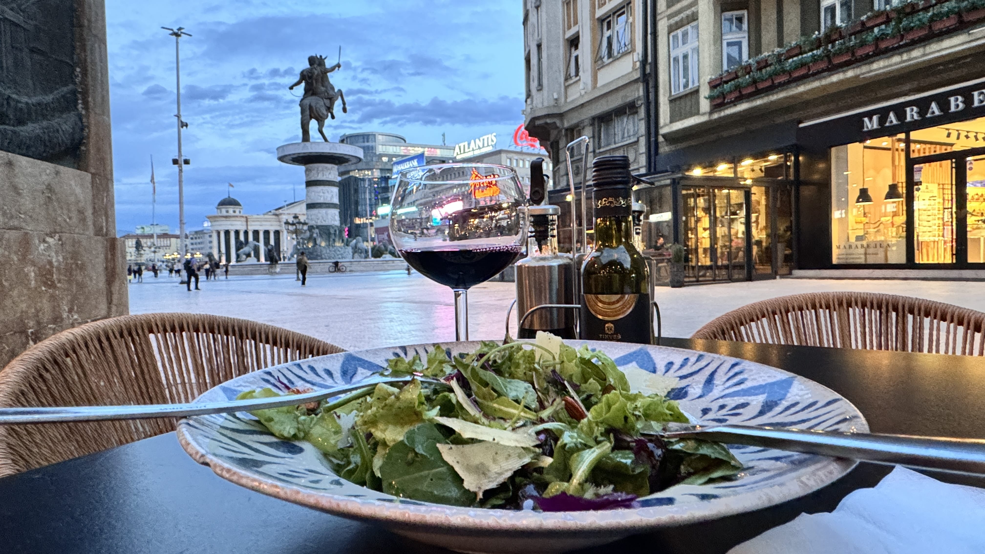 A close-up of a salad and glass of red wine on a café table at dusk in Skopje, North Macedonia, with the Warrior on a Horse statue and lit-up city buildings visible in the background.