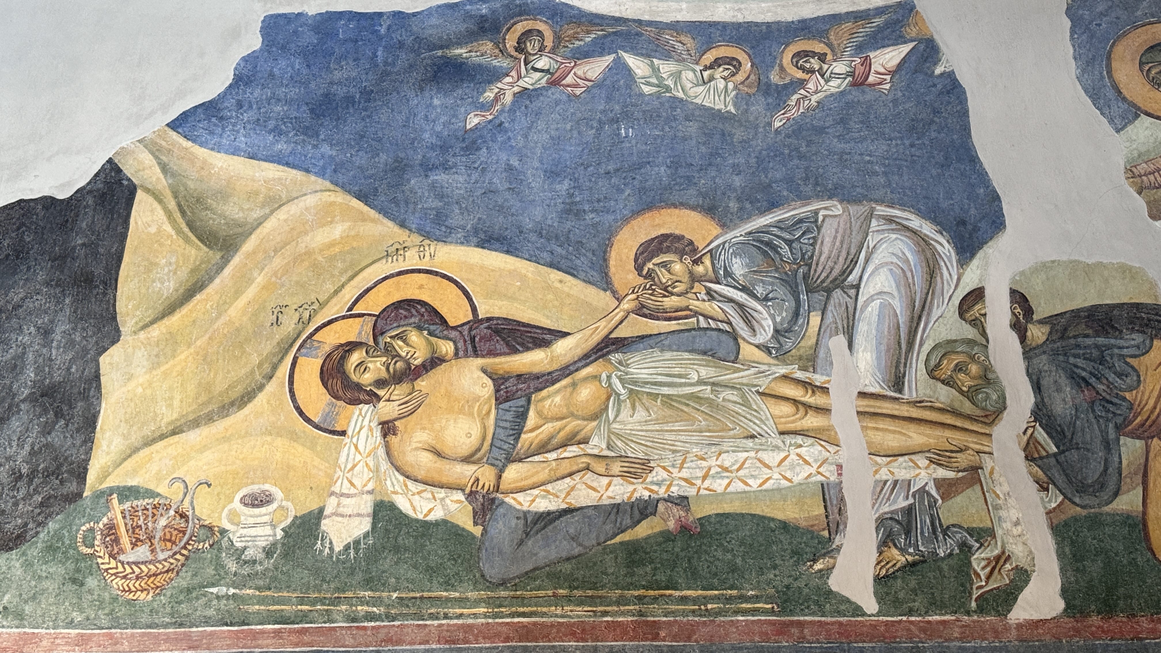 Fresco of the Lamentation of Christ, with Mary holding Jesus’ body after the crucifixion, surrounded by grieving figures in rich robes. The background shows angels and a dark blue sky, with part of the image faded or missing due to age.