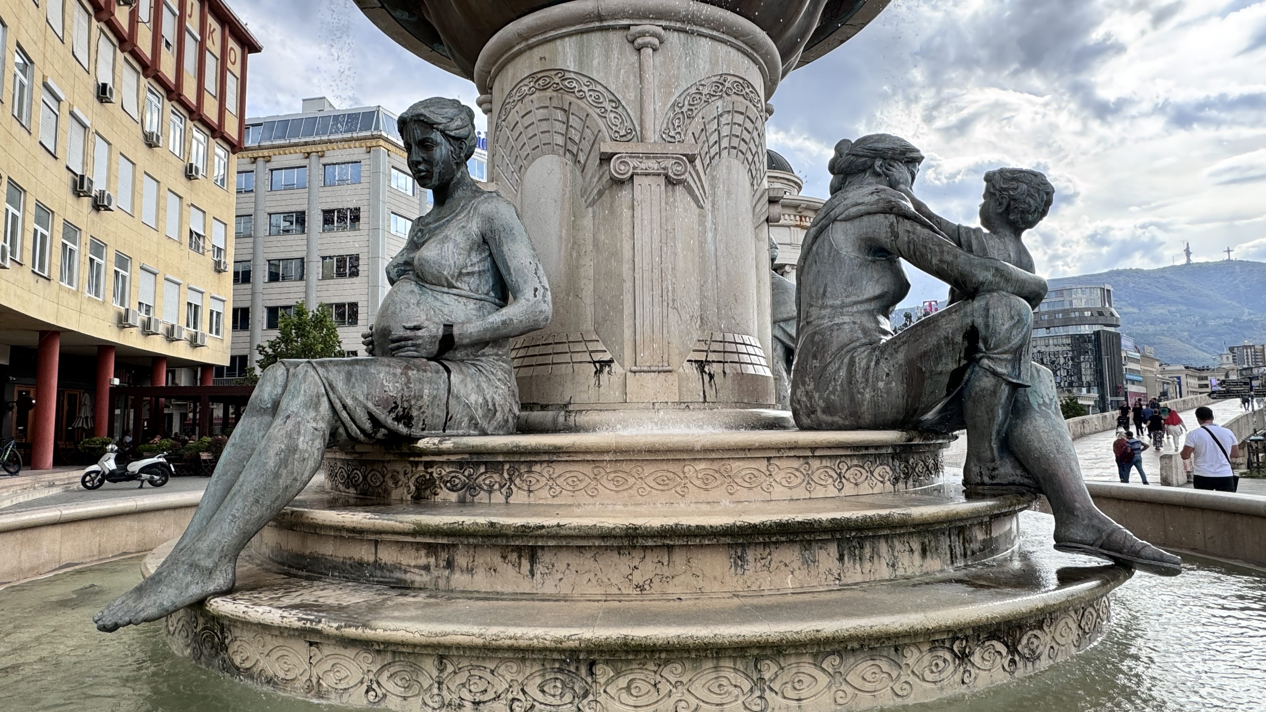 Bronze statues of two women—one pregnant, the other embracing a small child—seated at the base of a neoclassical-style fountain in Skopje. Buildings and a cloudy sky are visible in the background.