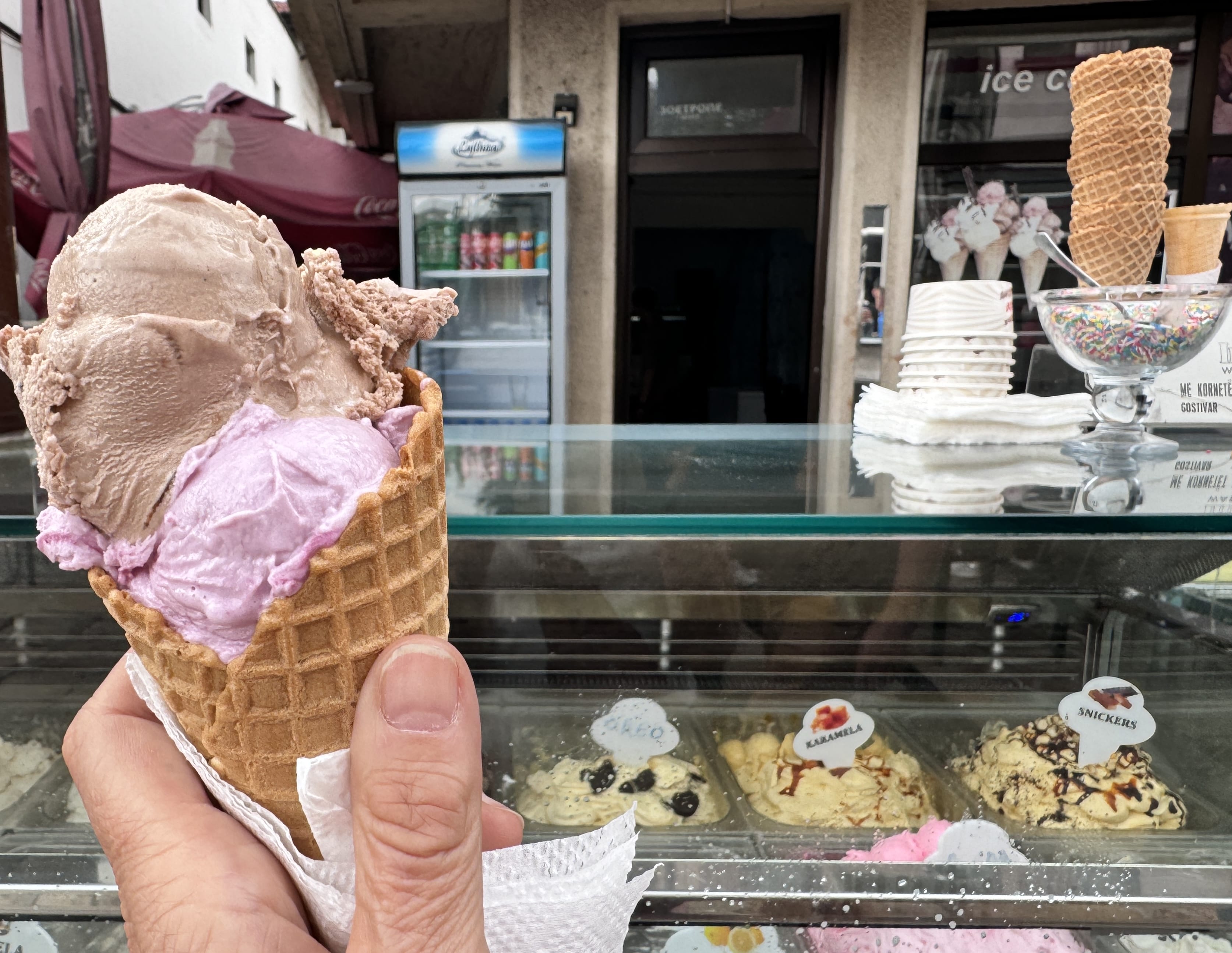 Hand holding a gelato cone with chocolate and blueberry scoops in front of a display case of ice cream flavors.