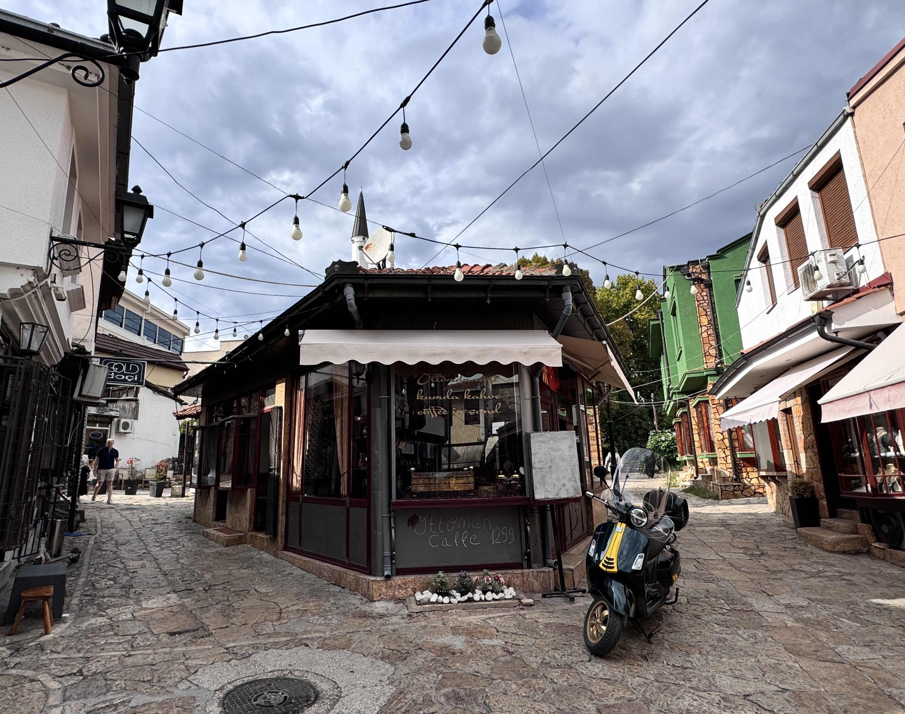 A small café on a cobblestone street in Skopje’s Old Bazaar, with string lights overhead, a scooter parked out front, and traditional buildings on either side.