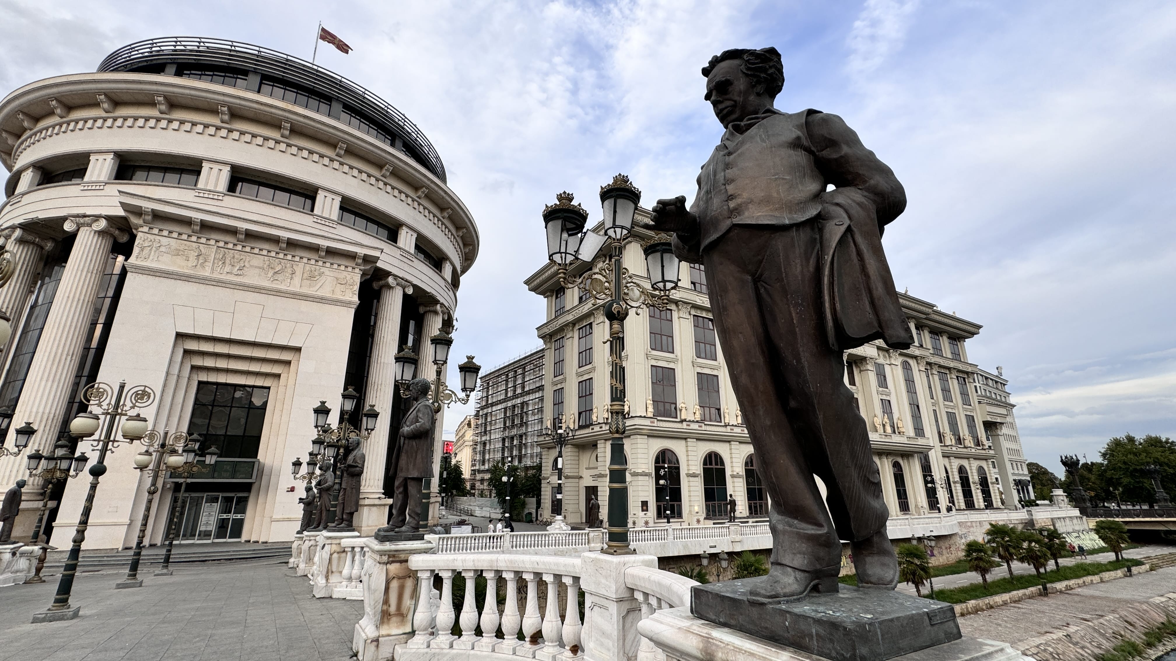 A row of bronze statues lines a bridge in Skopje, North Macedonia, with the nearest figure depicted in 19th-century attire. Behind it stands a grand neoclassical building with columns and reliefs, and more ornate facades are visible in the background under a cloudy sky.