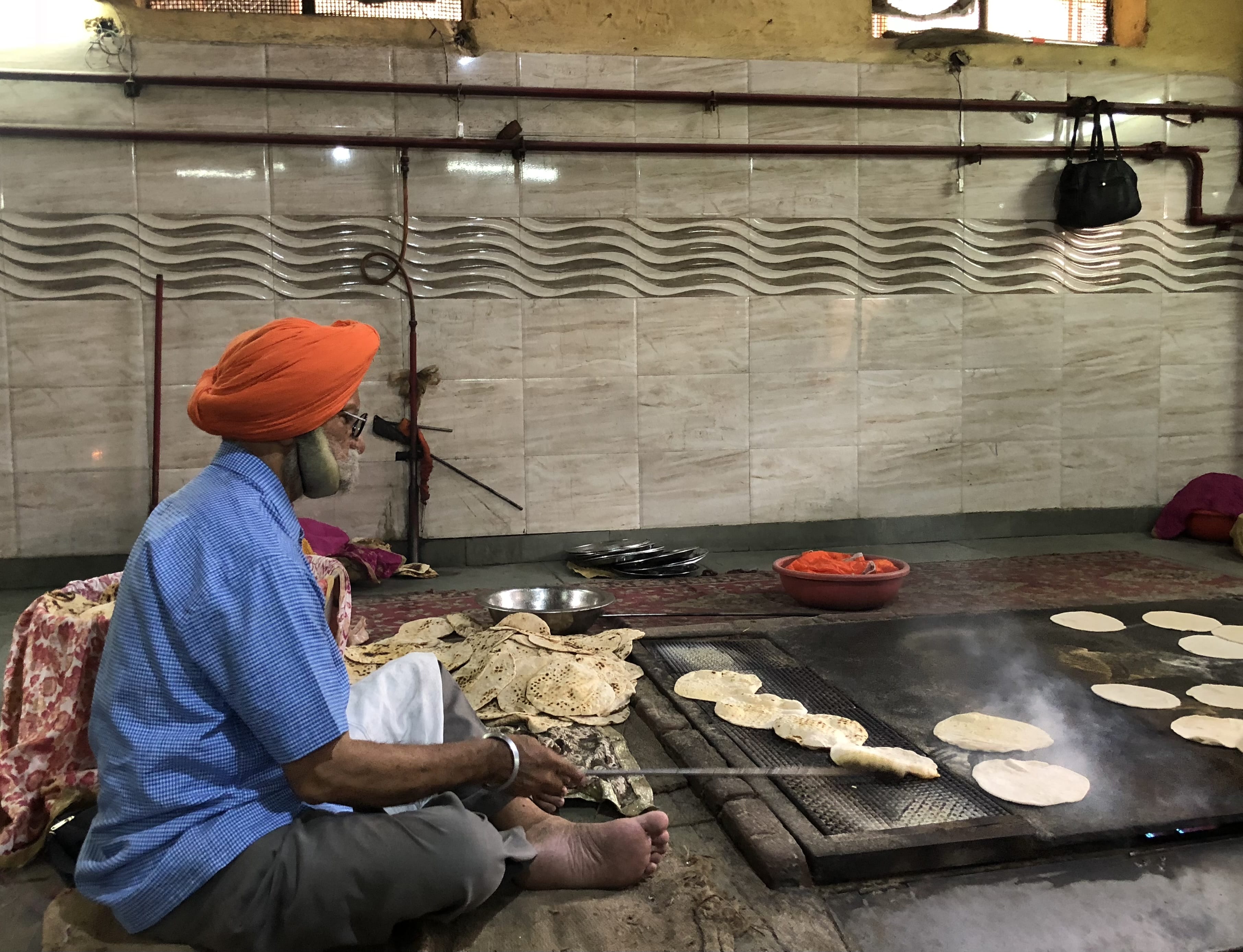Man in orange turban cooking rotis on a large griddle inside the community kitchen of Gurudwara Bangla Sahib.