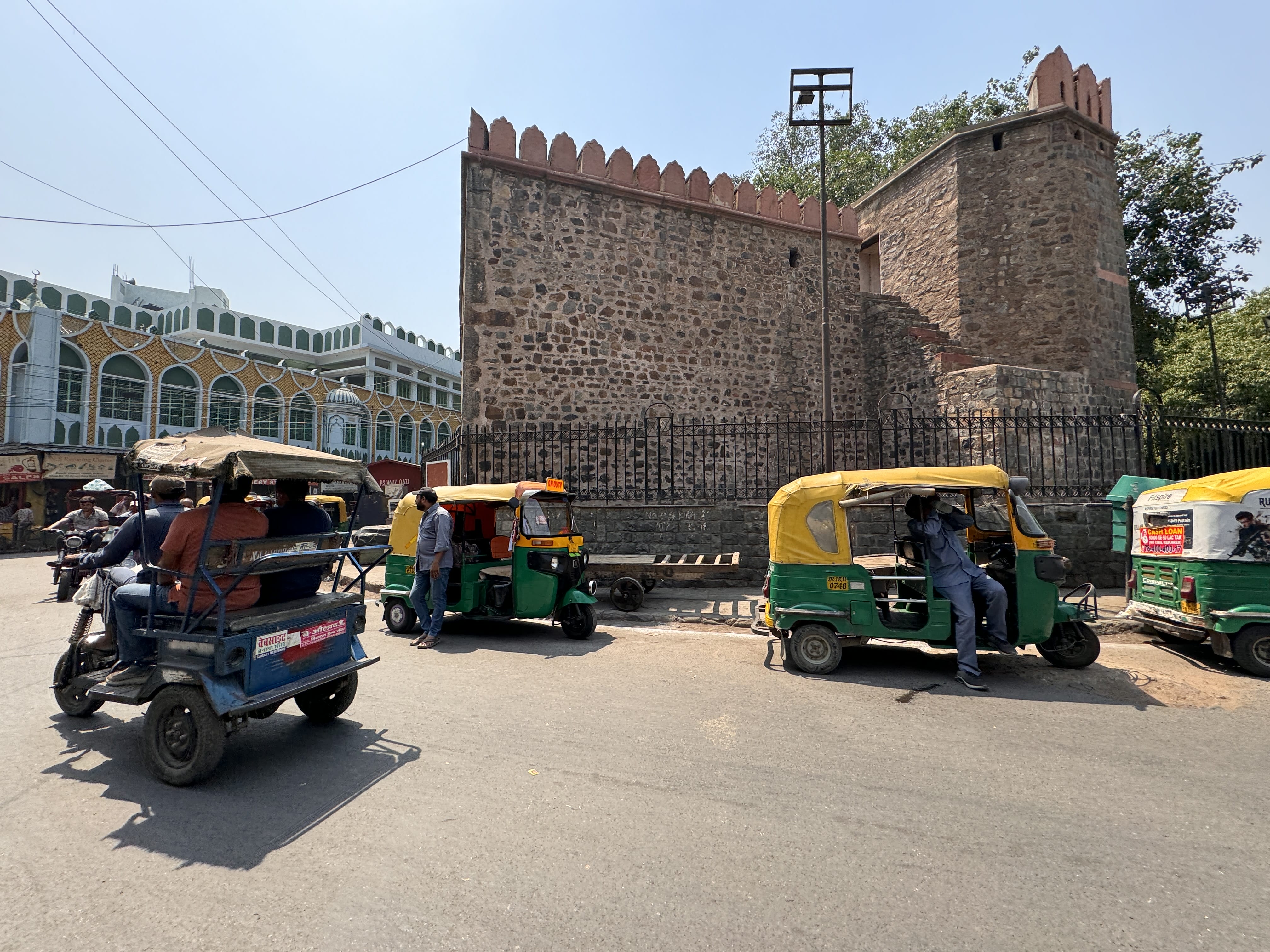 Ajmeri Gate, a historic stone gateway in Old Delhi, stands tall with red crenellations and weathered walls. Auto rickshaws and people pass by in the foreground.