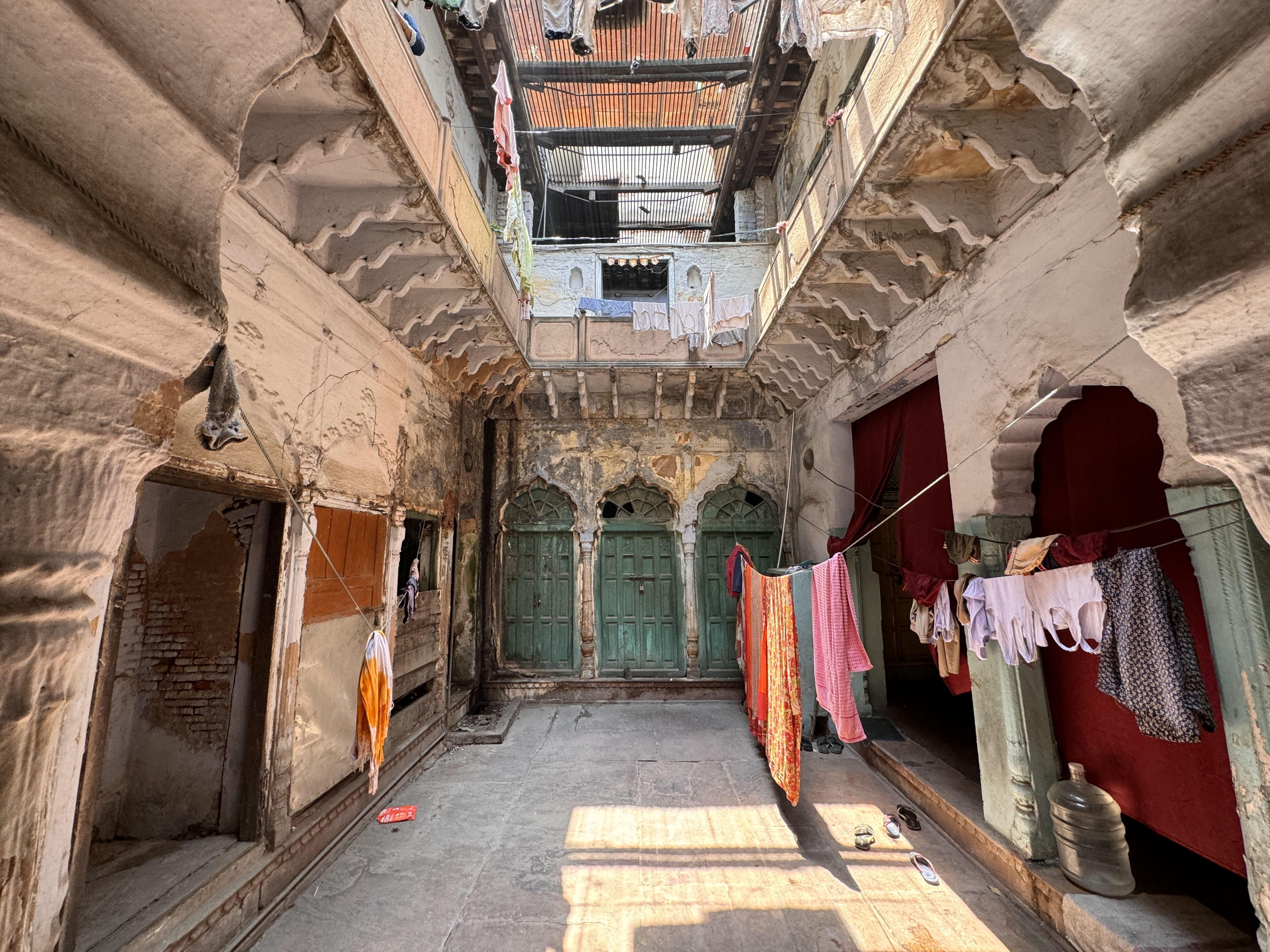 Interior courtyard of an old haveli with faded green doors, crumbling plaster, and colorful saris hanging on a clothesline across the space.
