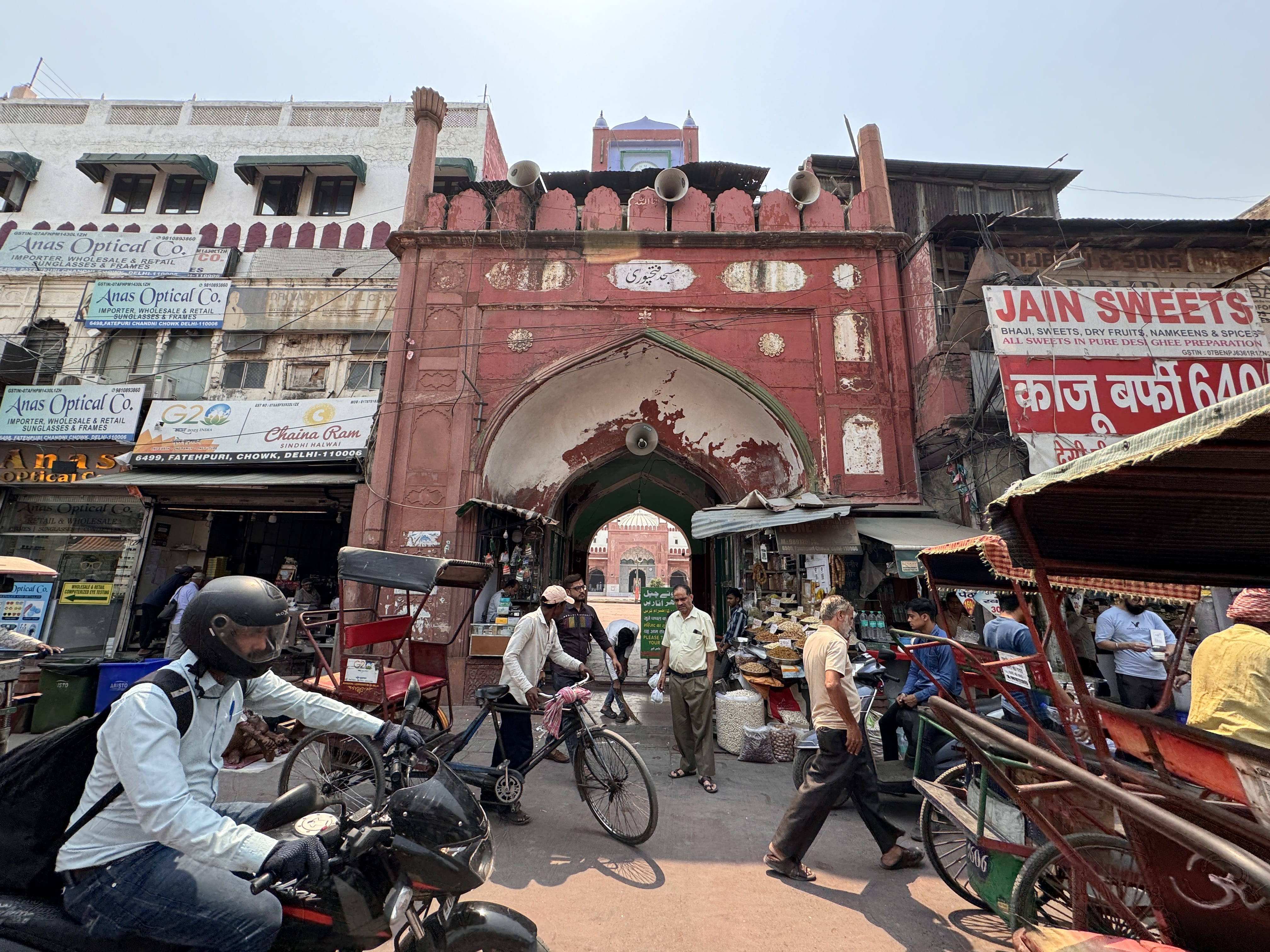 Crowds passing through a large red Mughal-style gateway in Old Delhi, surrounded by shops and market chaos.
