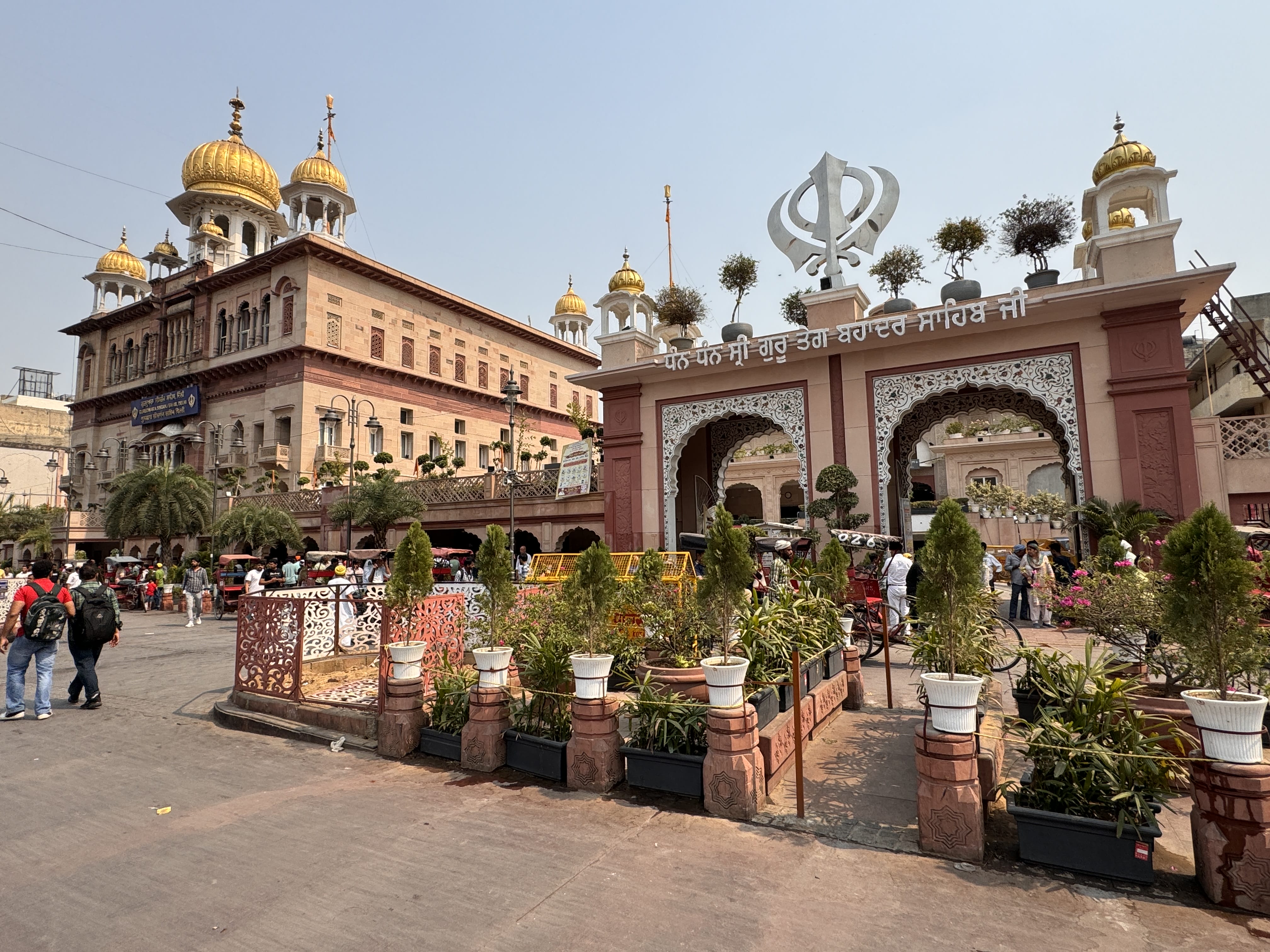 Visitors and rickshaws outside the grand arched entrance to Gurudwara Bangla Sahib in Delhi, with golden domes and decorative plants in the foreground.