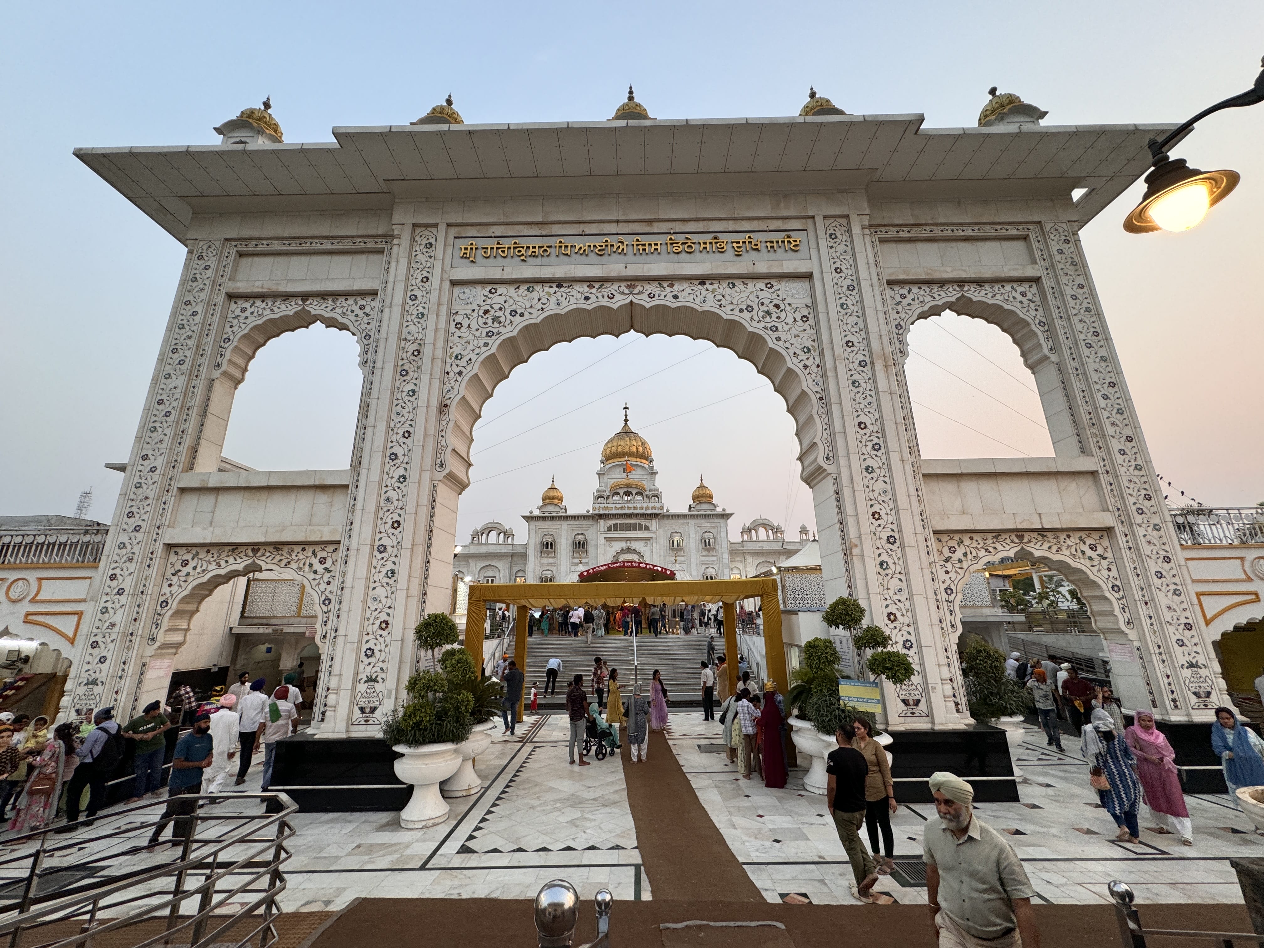 Ornate white marble gateway leading to Gurudwara Bangla Sahib in Delhi, with golden domes visible in the background and visitors walking through the entrance.