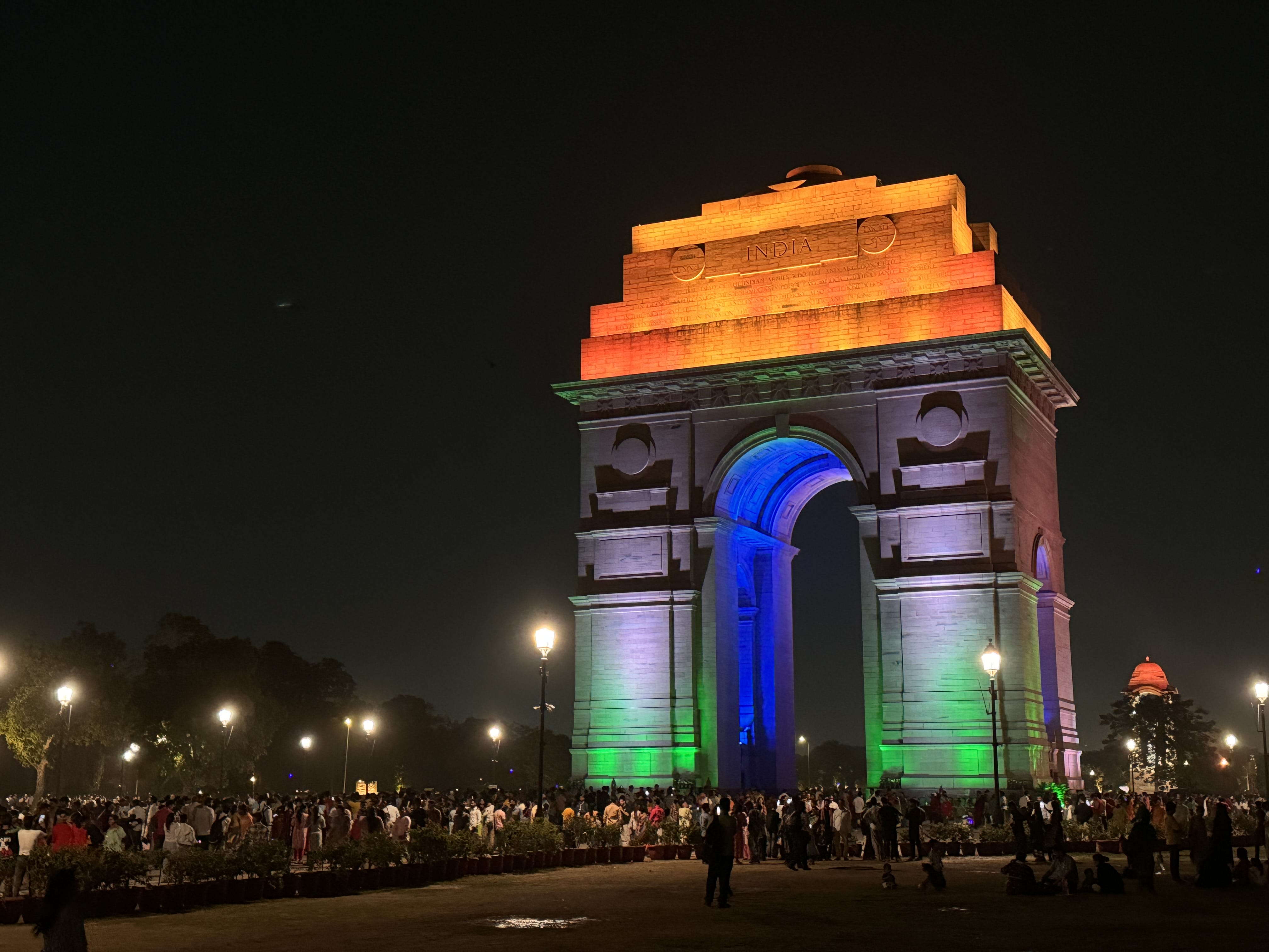 India Gate illuminated in orange, white, green, and blue lights at night, with a crowd gathered around the base.