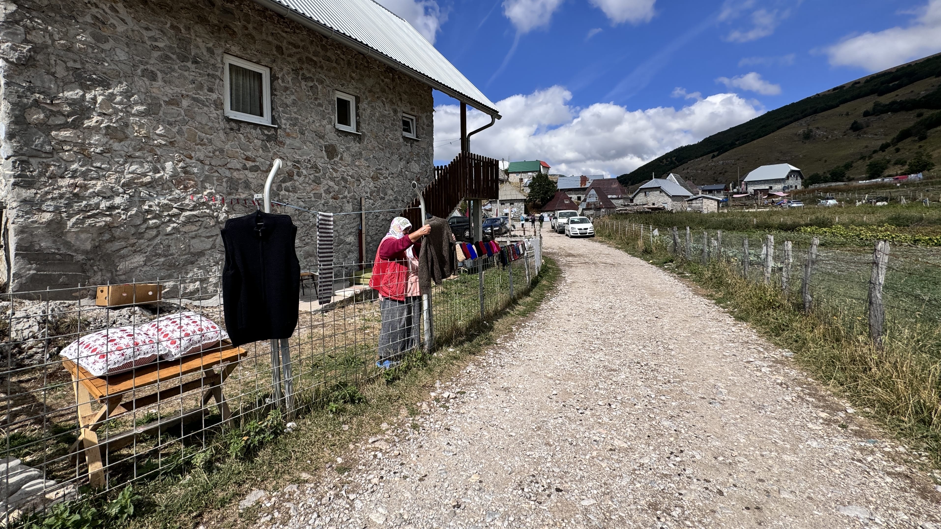 Elderly woman hanging handmade knitwear on a fence outside a stone house in Lukomir village, Bosnia and Herzegovina.