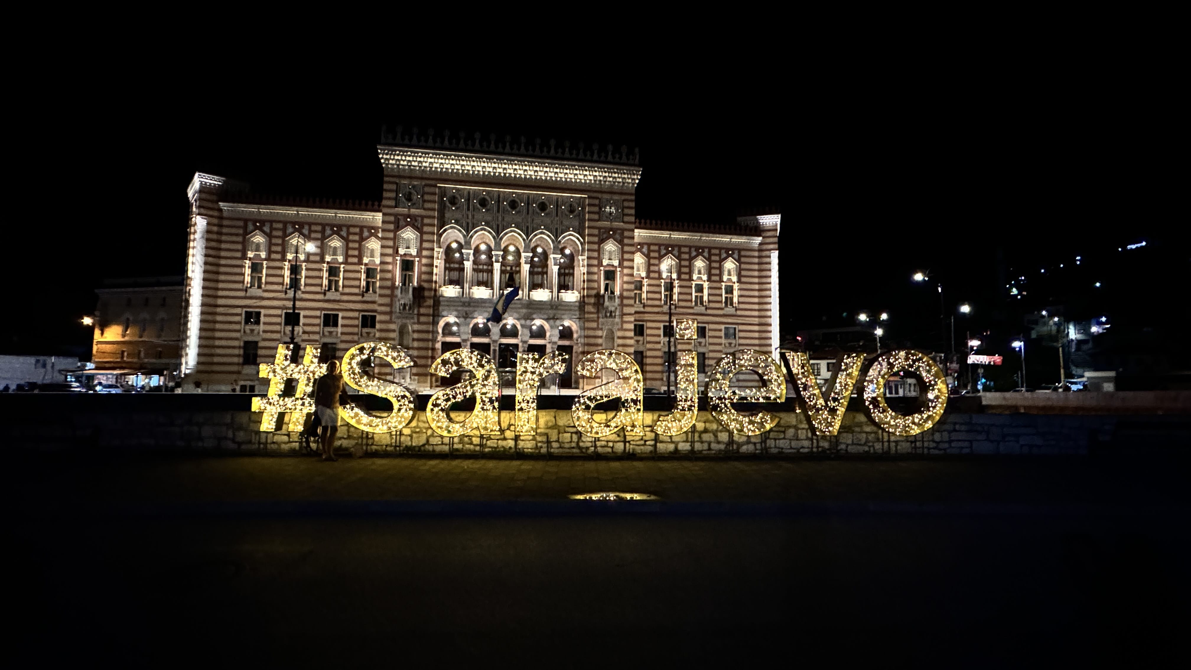 Illuminated #Sarajevo sign in front of the historic Vijećnica (City Hall) building at night.