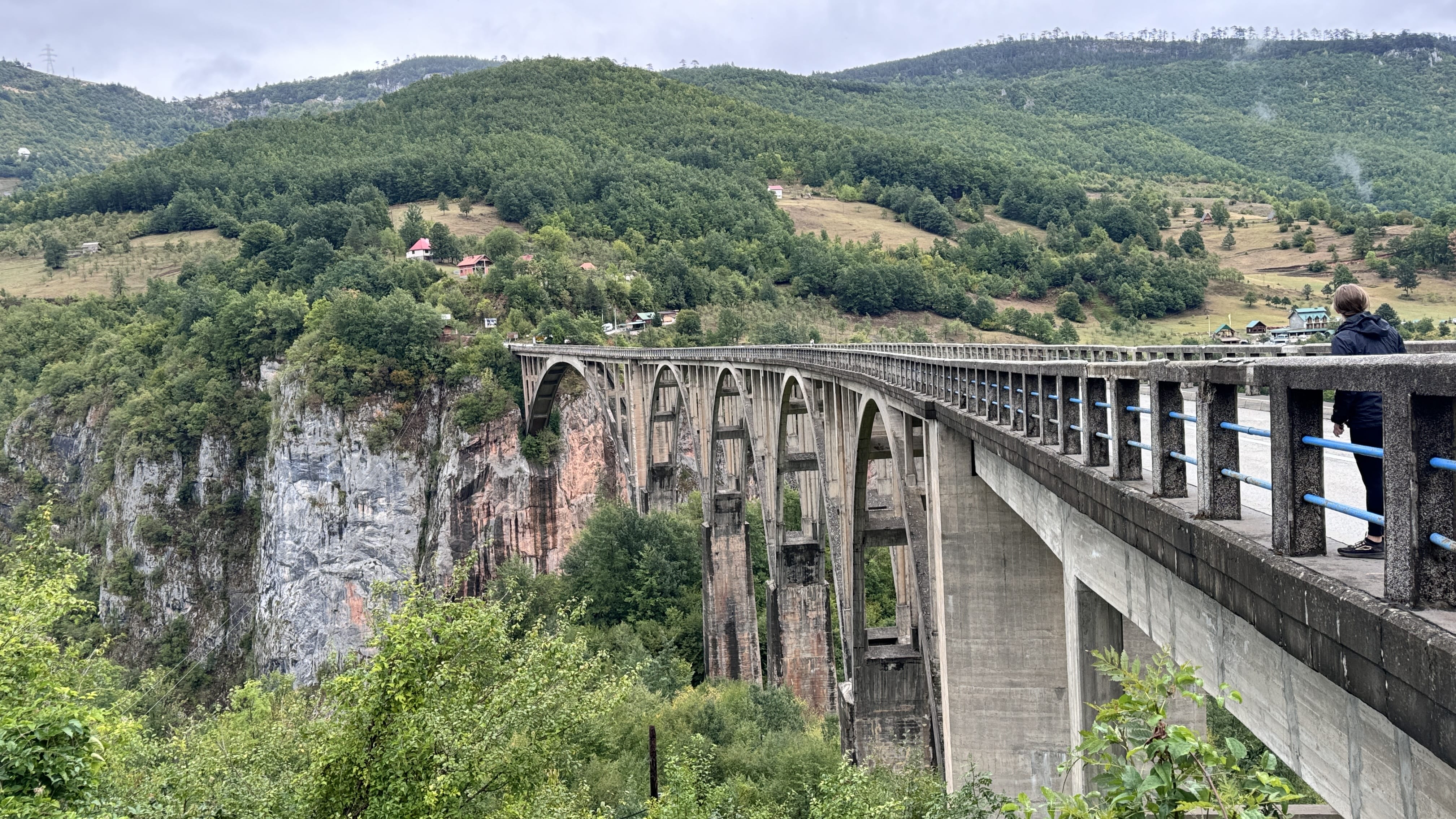 Concrete Tara Bridge stretching over a deep canyon in Montenegro with forested hills and scattered red-roofed homes in the background.