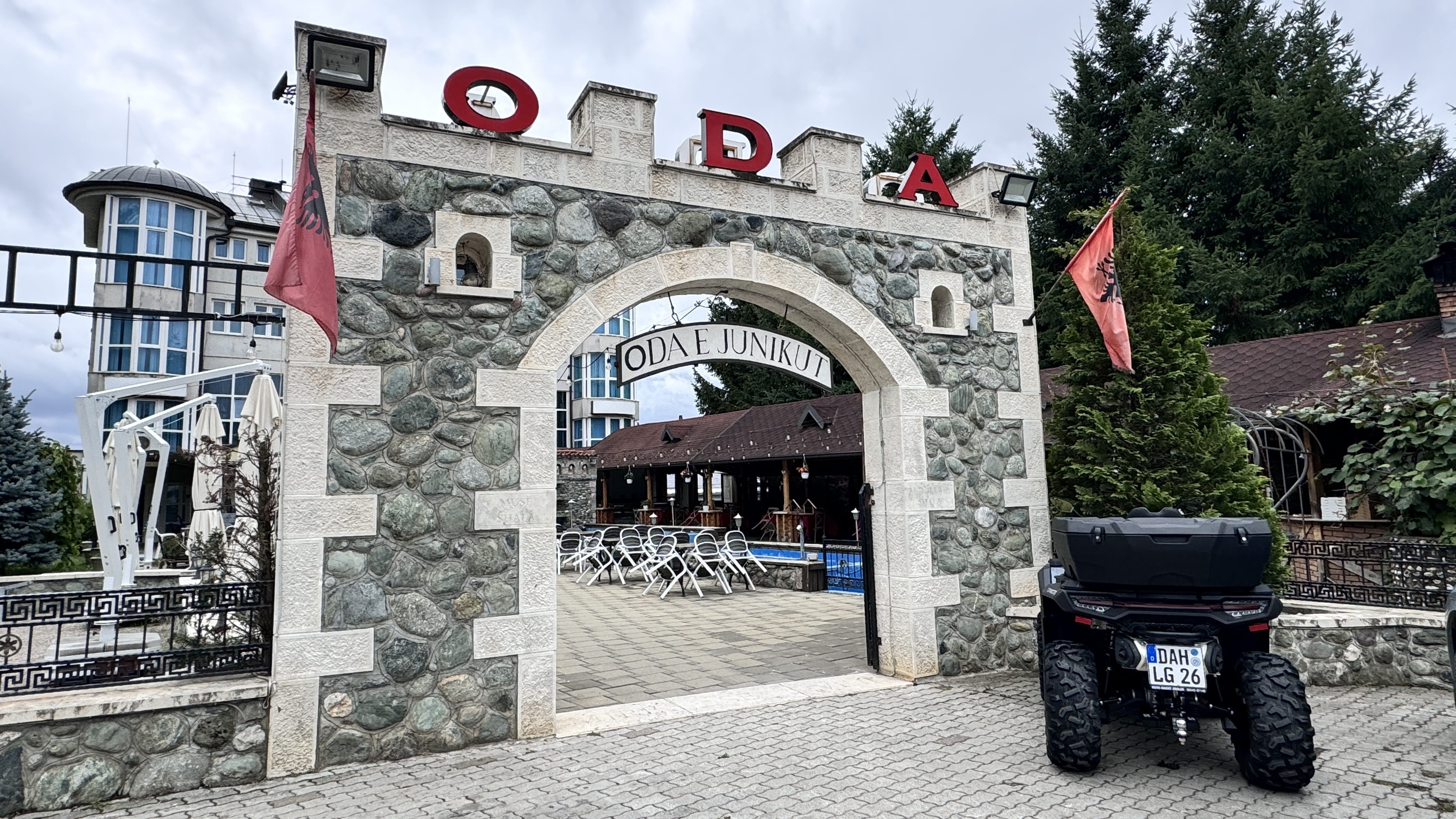 Stone archway entrance to the traditional-style restaurant "Oda e Junikut" in Kosovo, with Albanian flags and an ATV parked outside.